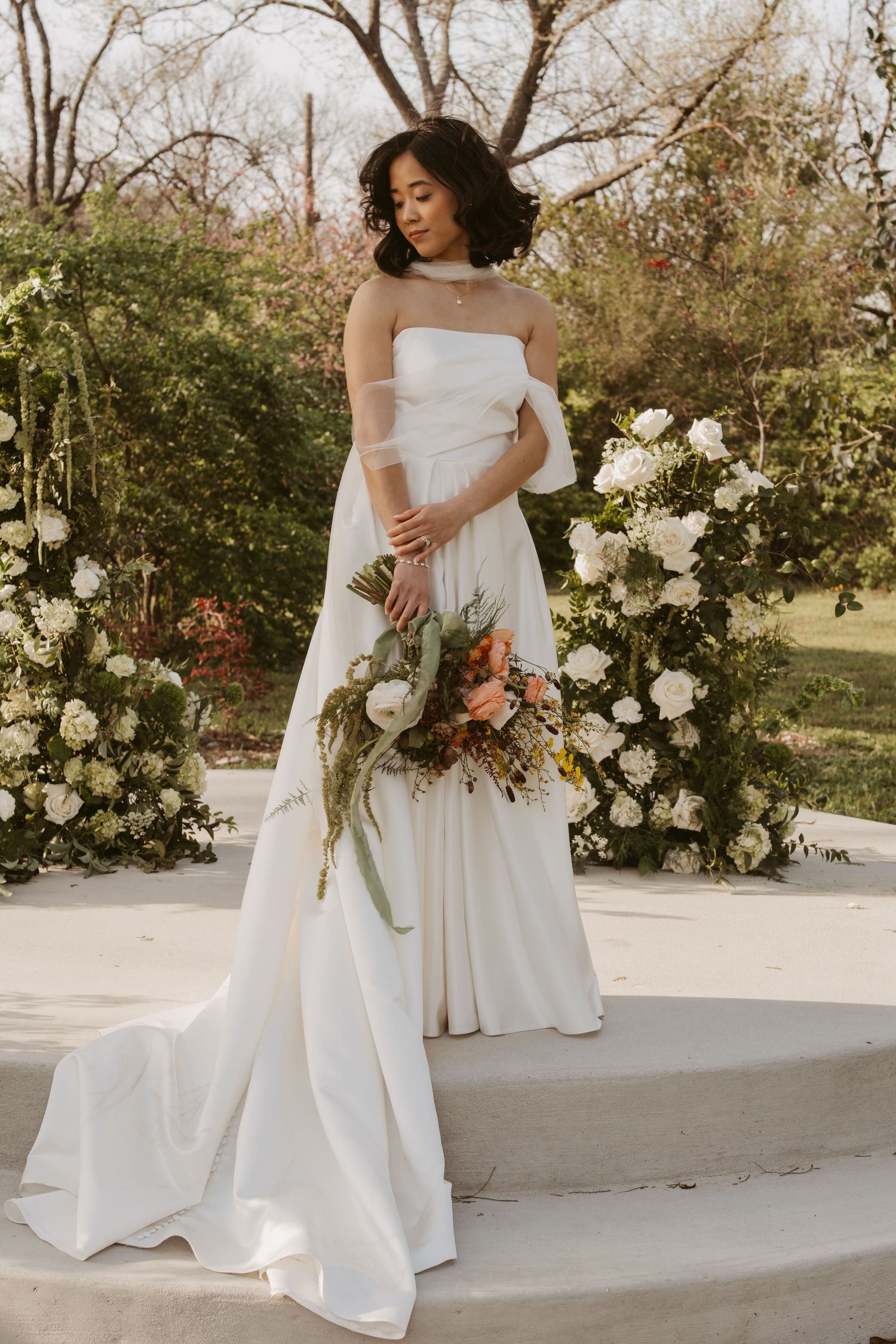 Bride in strapless white satin gown holding floral bouquet stands on steps beside white rose arrangements outdoors.