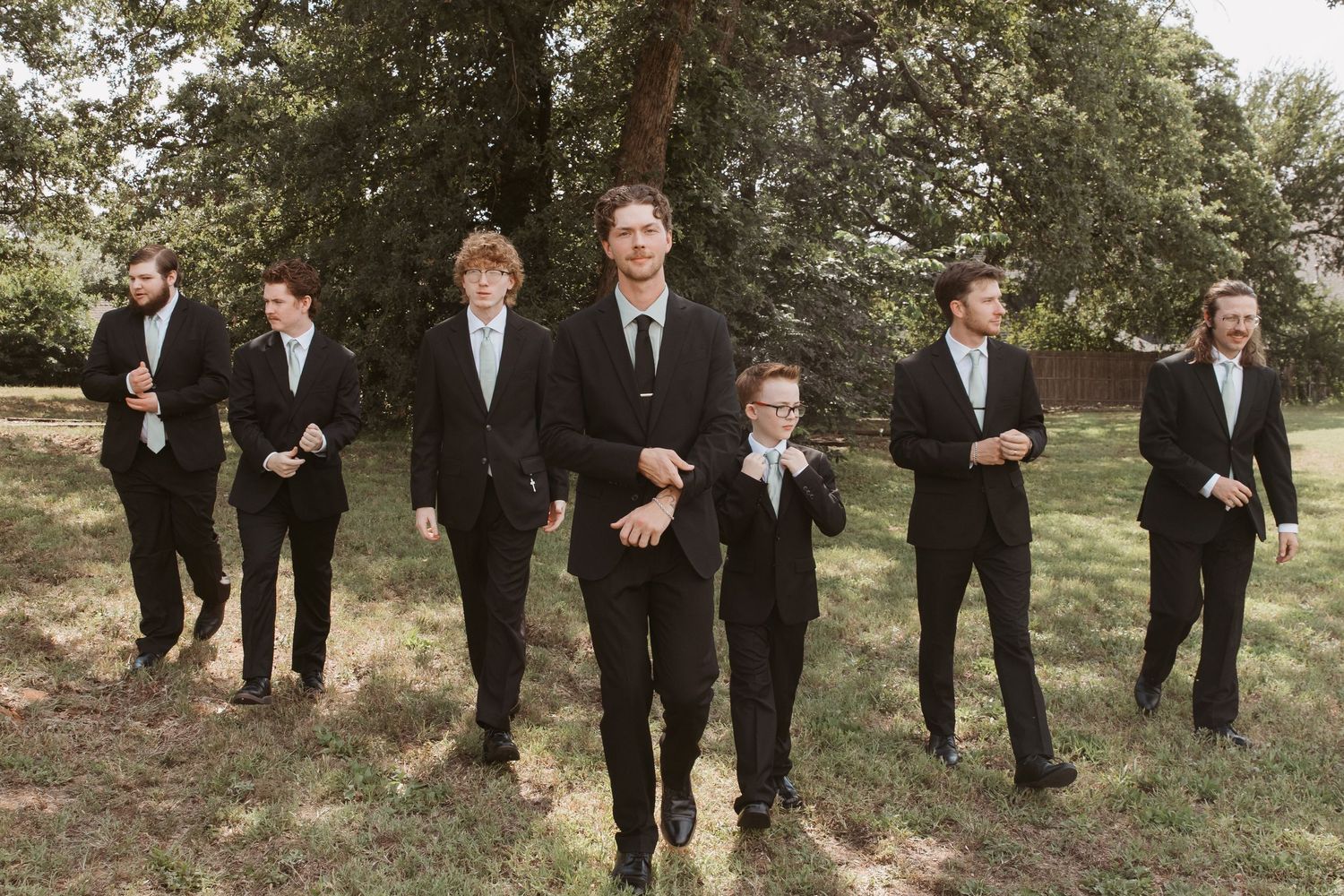 Groomsmen in black suits walking outdoors on grass with trees in background at a wedding.