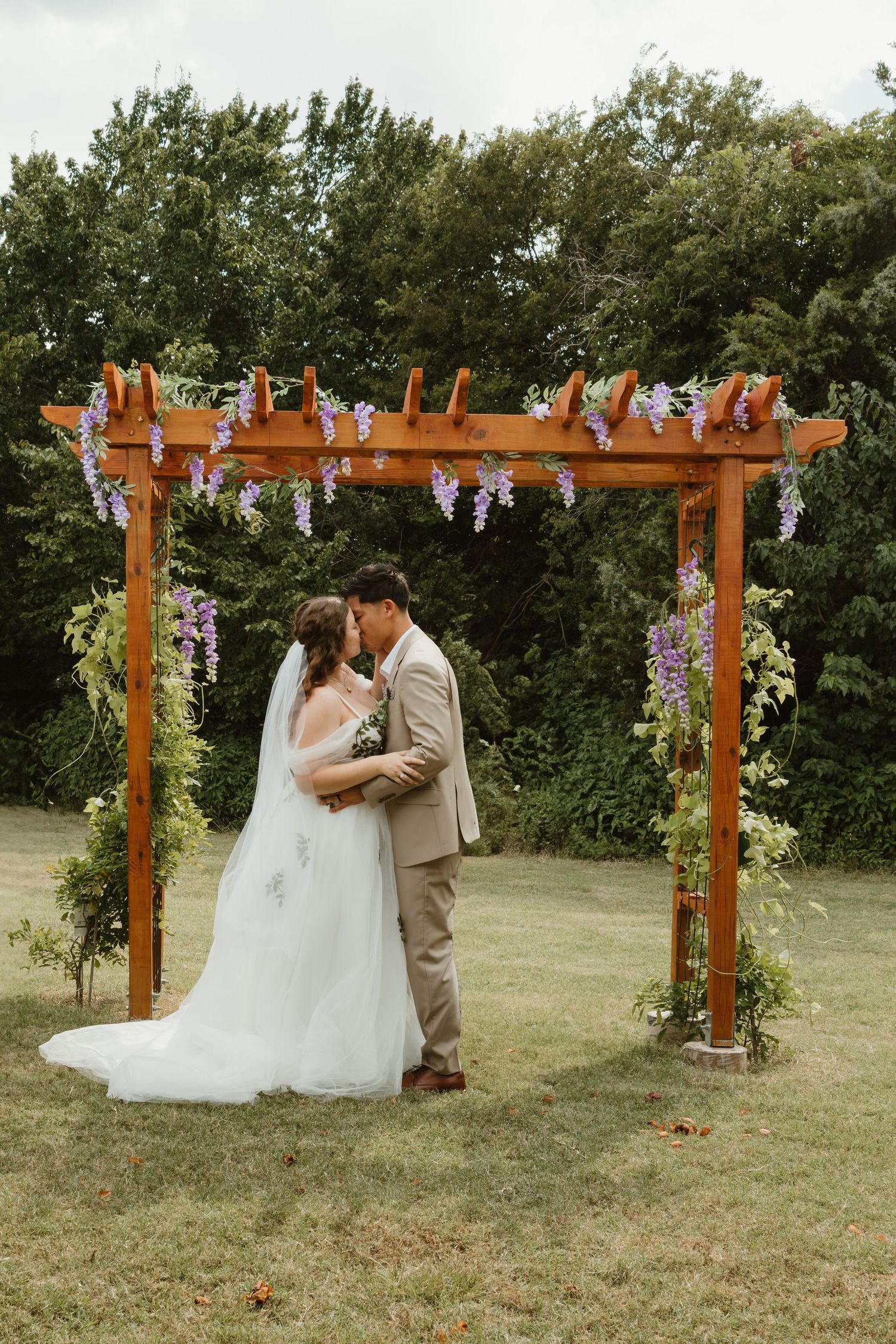 Wooden pergola decorated with purple wisteria flowers creates romantic backdrop for wedding ceremony in garden setting.