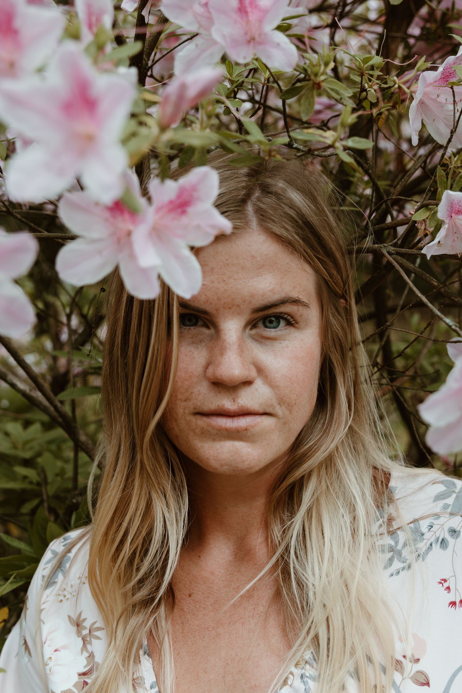 Portrait of a woman with blonde hair surrounded by blooming pink azalea flowers in a garden setting.