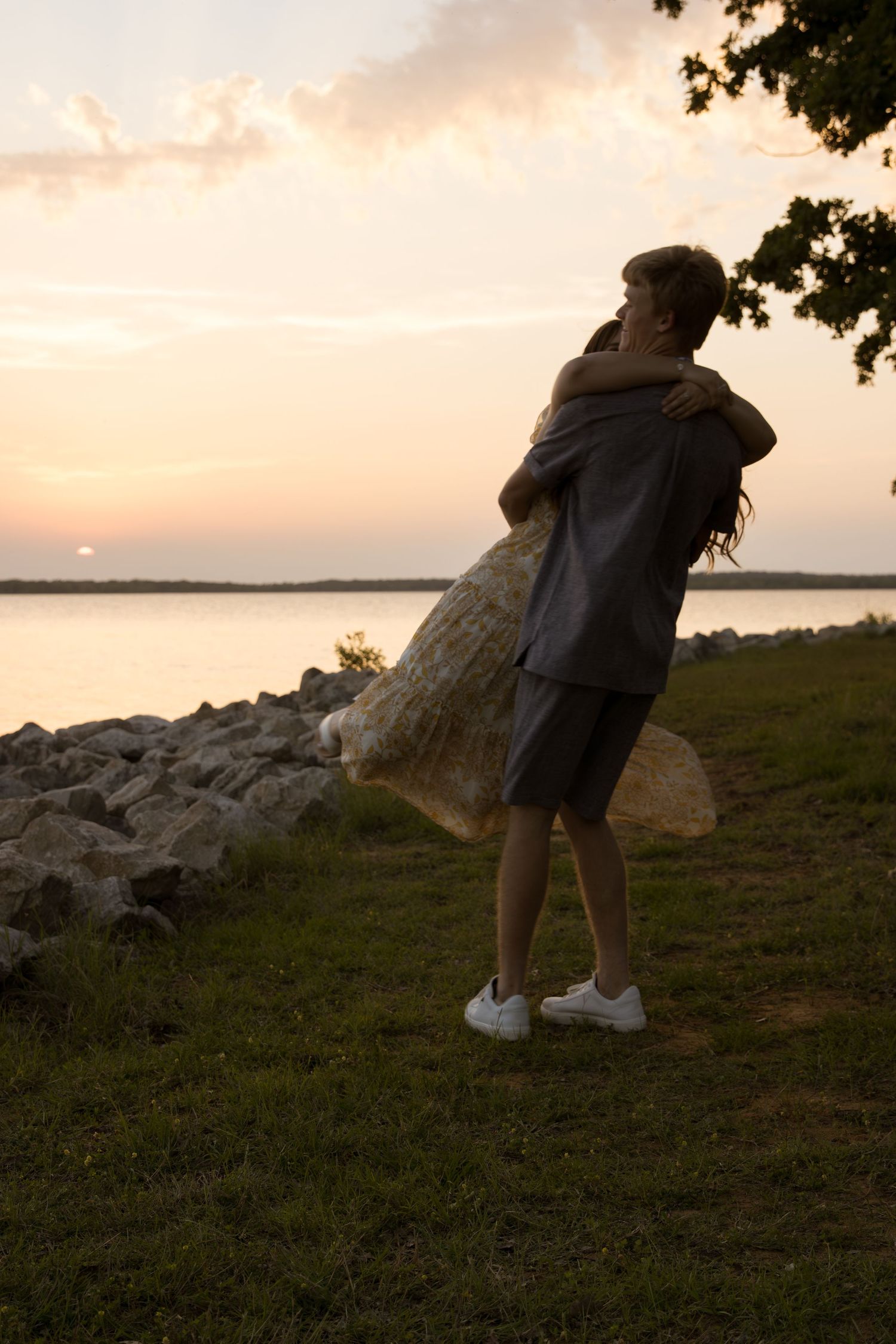 Silhouetted figure stands on rocky shoreline watching sunset over calm lake waters on a peaceful evening.