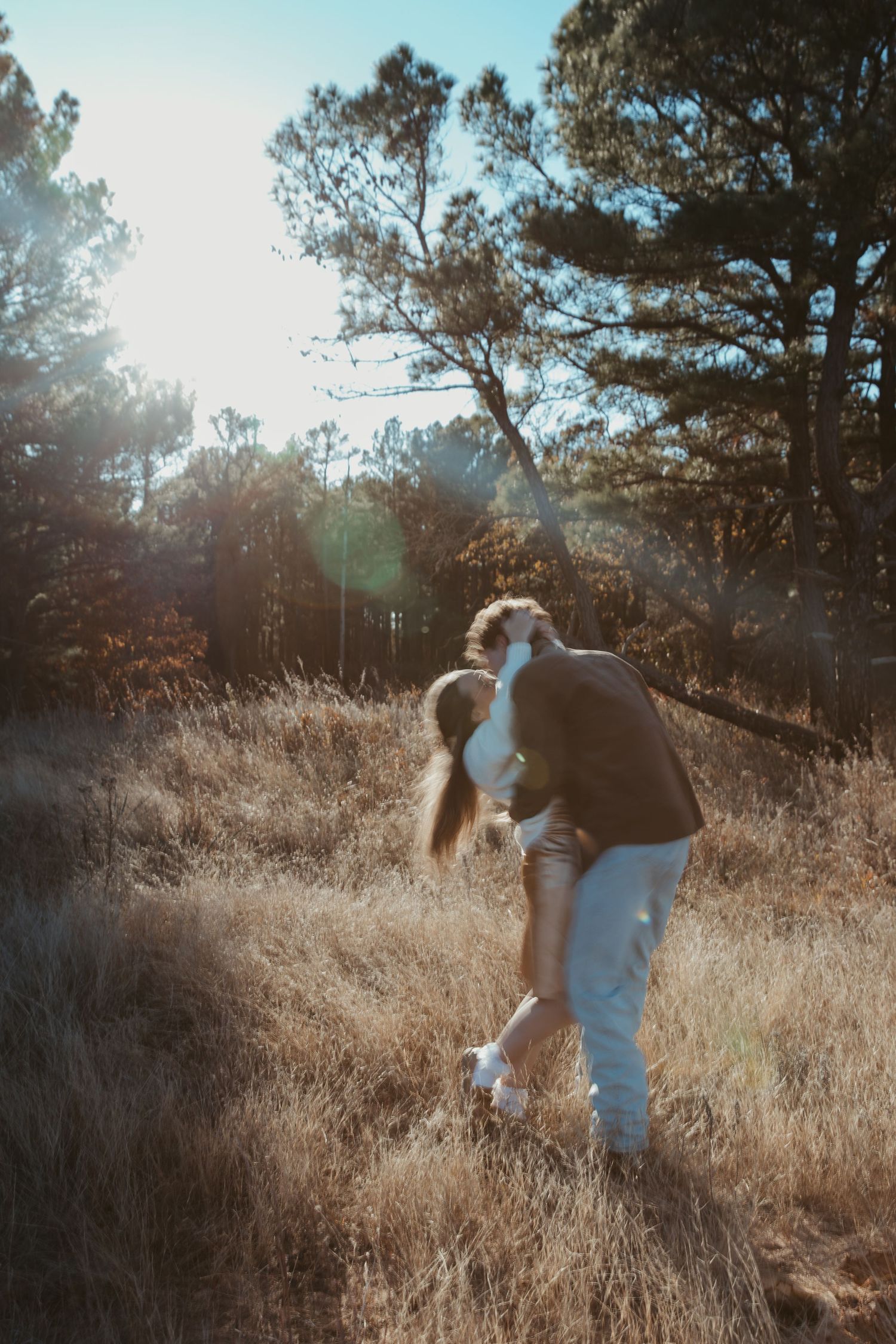 A couple embraces in a sun-drenched field with tall grass and trees creating a romantic outdoor setting engagement photos DFW
