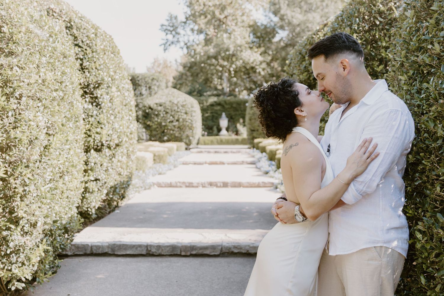 Romantic couple shares tender moments in a sunlit garden pathway surrounded by lush greenery and stone walls.