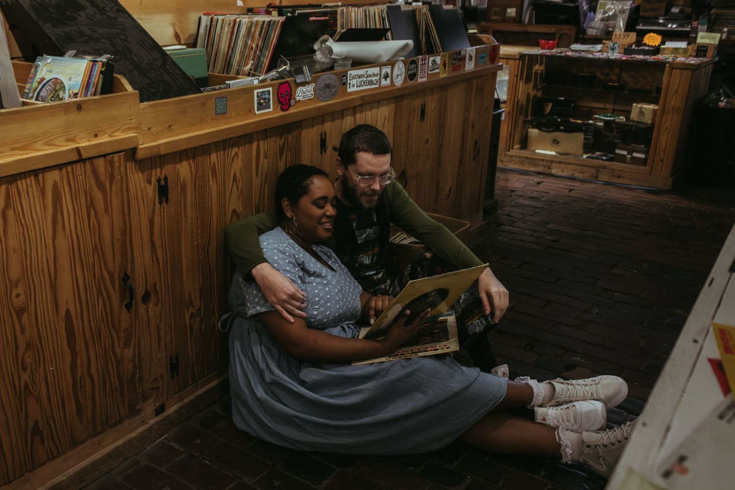 A cozy moment shared between two people sitting on the floor against wooden paneled walls in a dimly lit room.