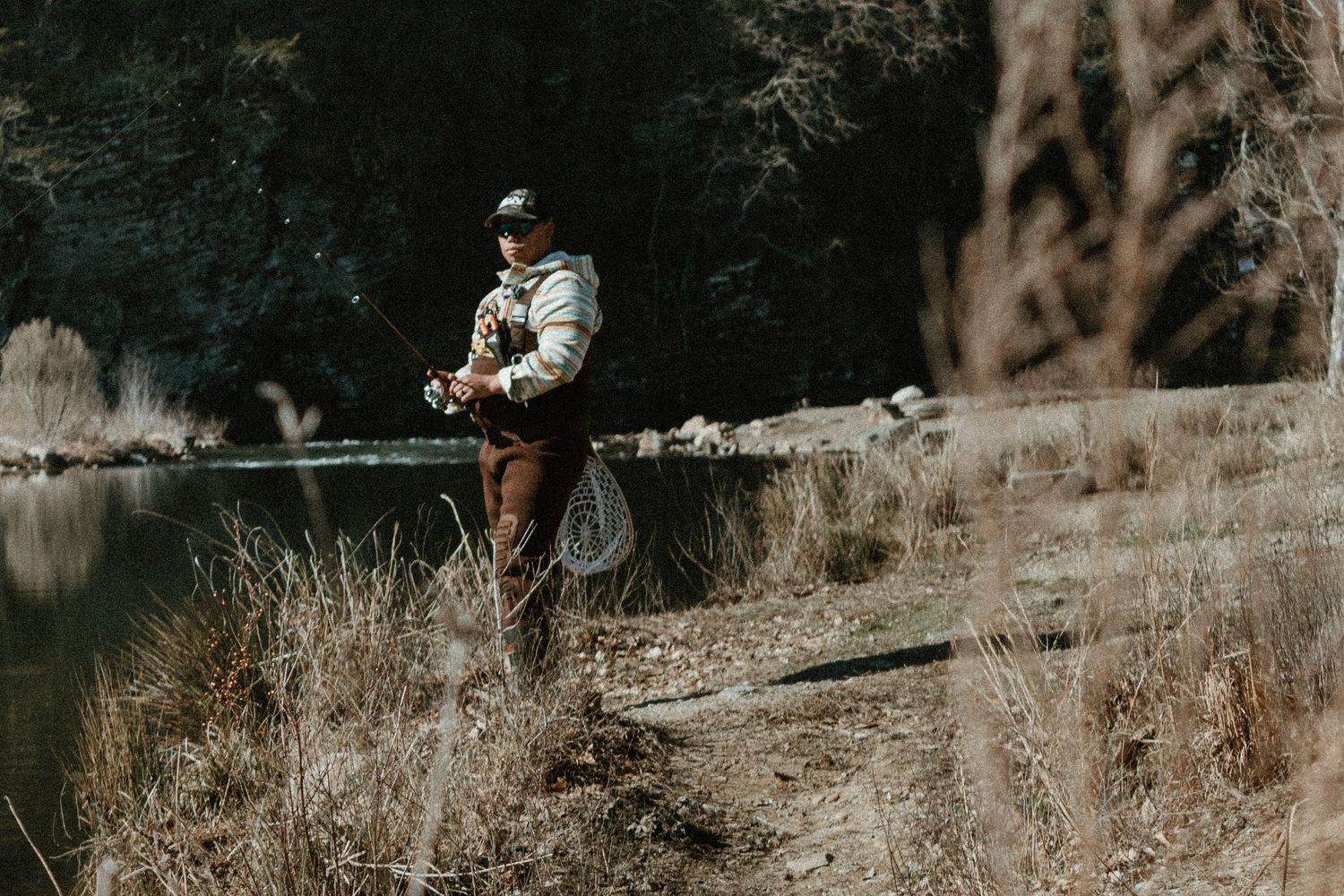 A fisherman stands on the riverbank casting a line during an overcast day.