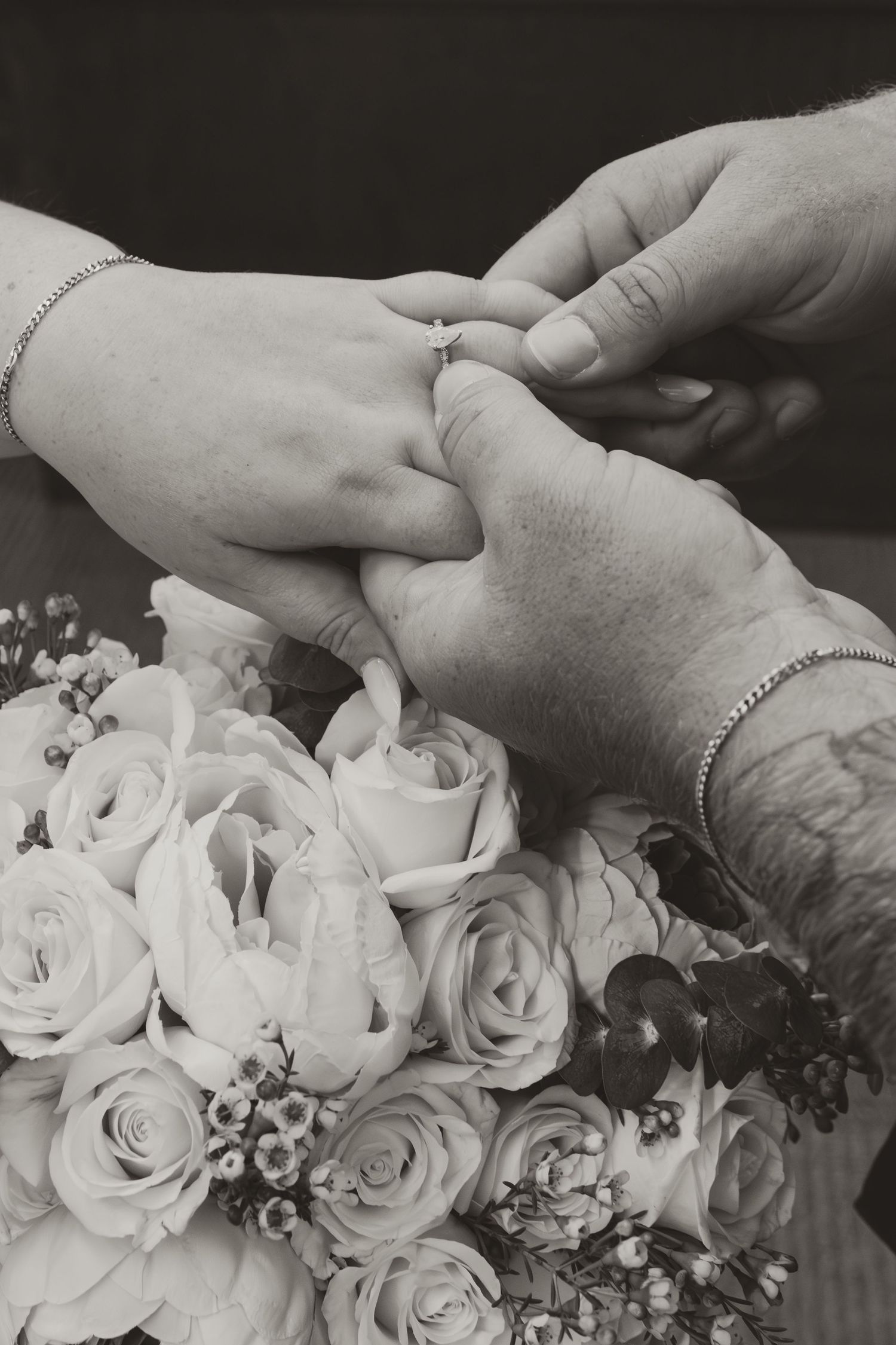 A black and white photograph shows hands placing a ring with a bouquet of white roses in the background.