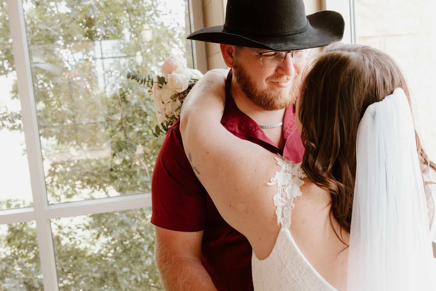 An intimate wedding moment near sunlit windows with burgundy and white attire.