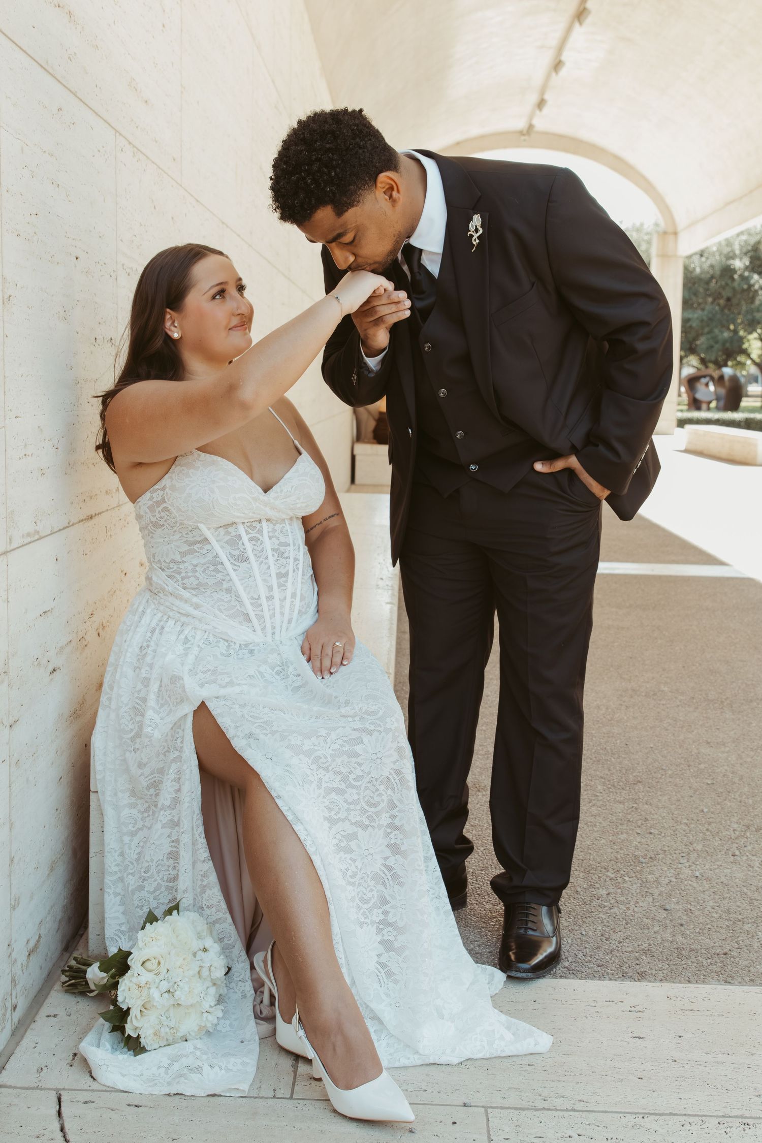 A wedding couple poses playfully near white stone columns, with the bride showing off her dress and shoes. Wedding Photographer DFW