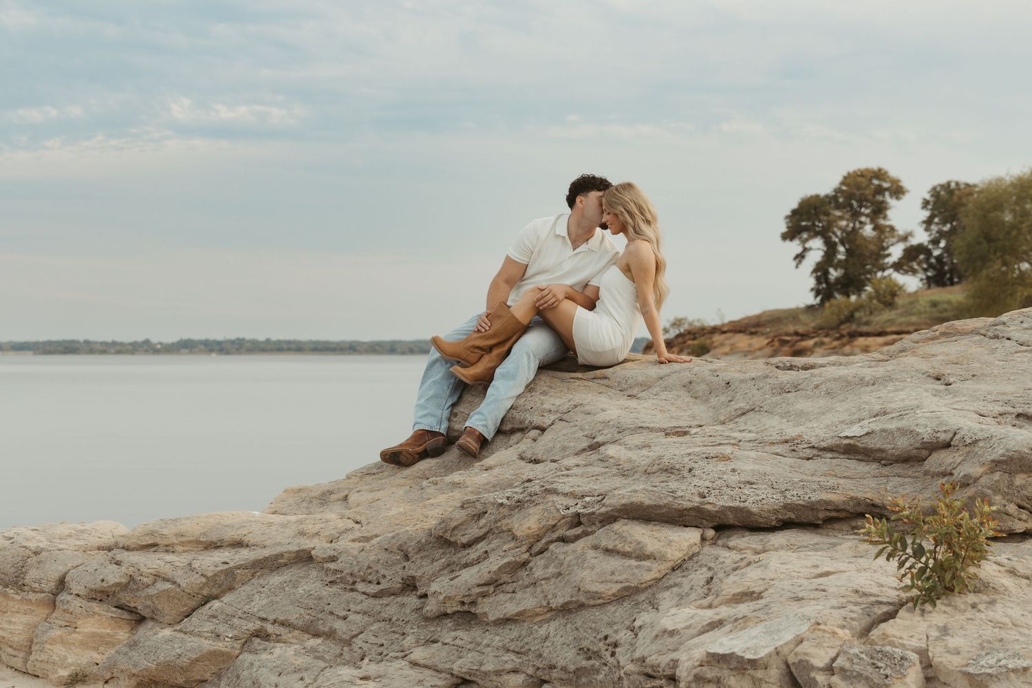 A couple sits together on rocky outcrop overlooking a serene lake during golden hour.