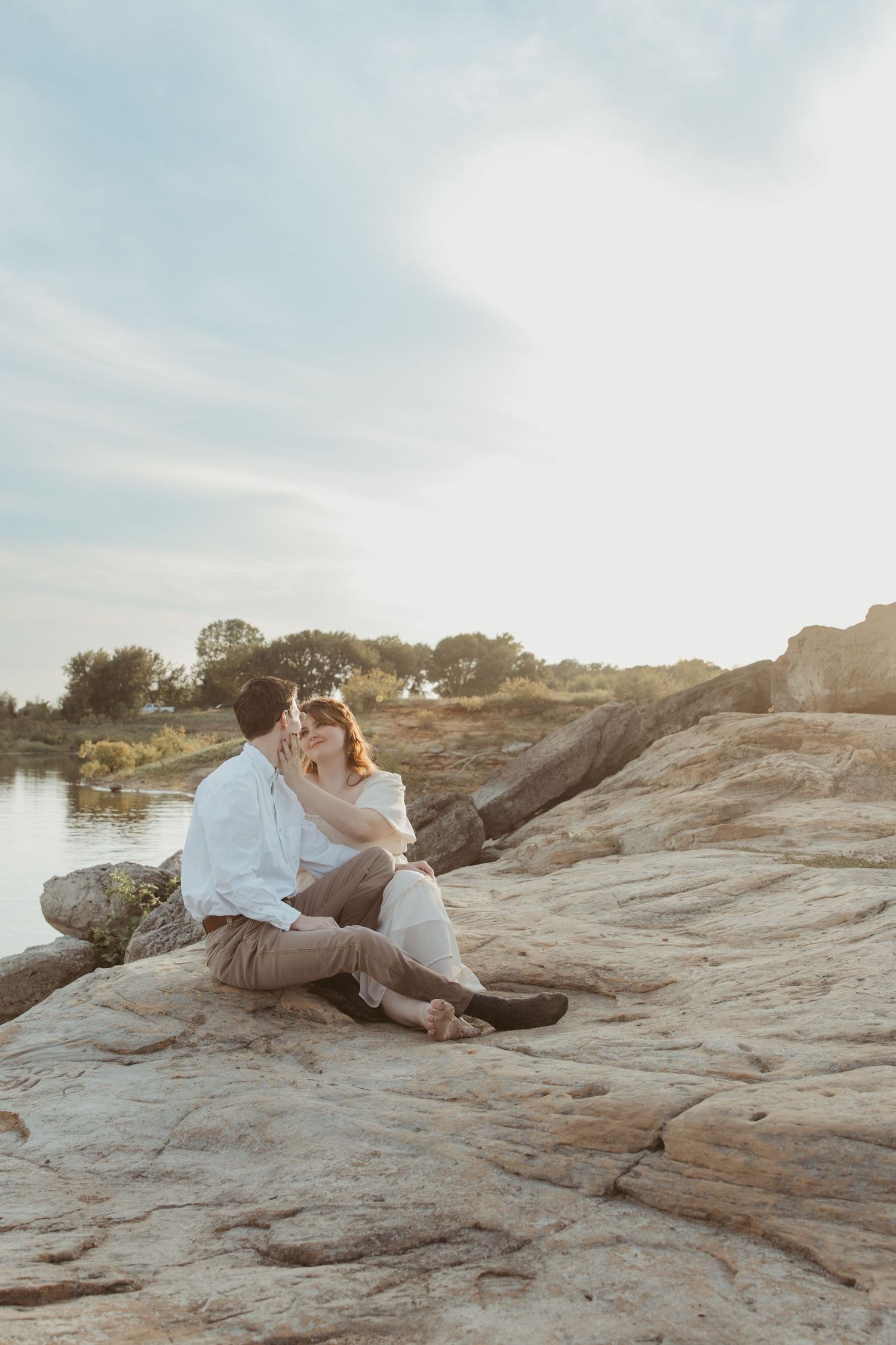 A couple sits close together on sun-warmed rocks by a peaceful lake during golden hour.