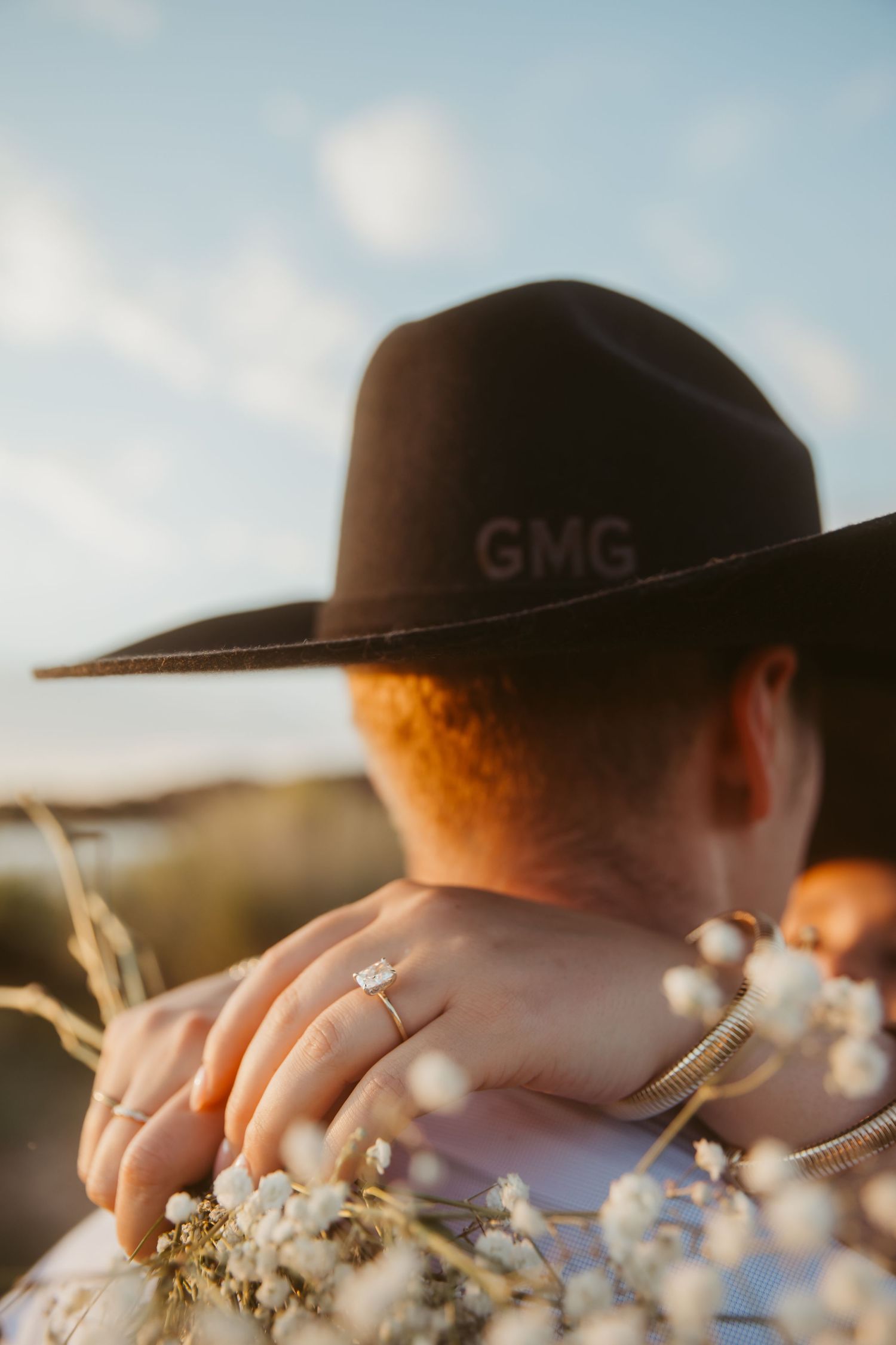 Close-up of an engagement ring on a woman's hand resting on a man's shoulder wearing a GMG cowboy hat.