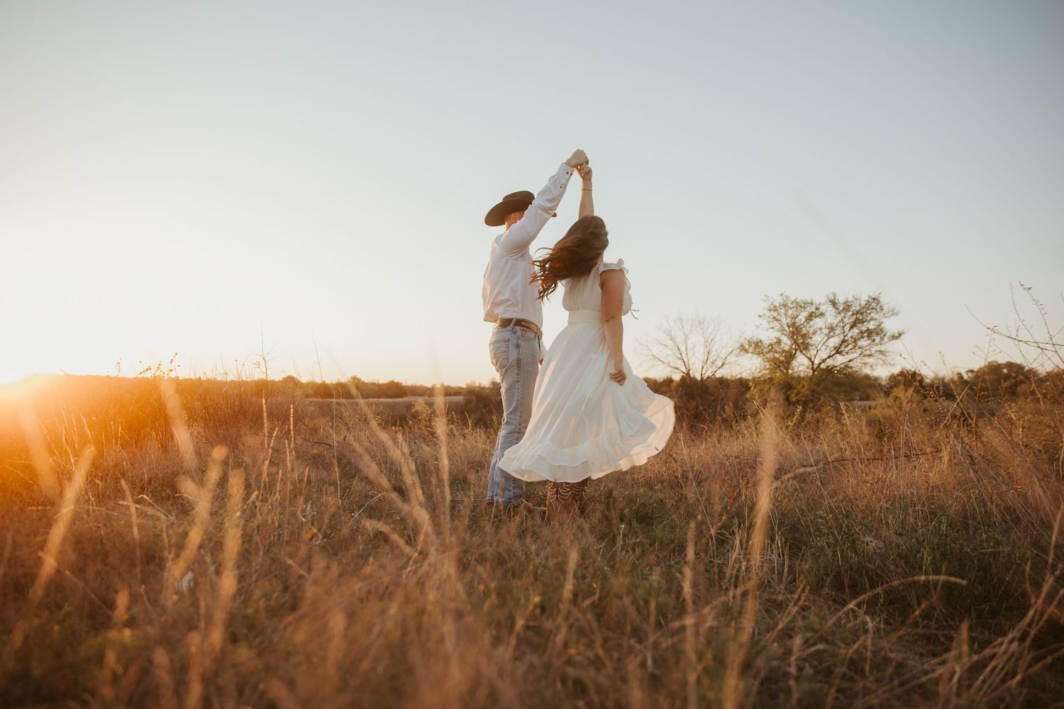 Couple dancing in golden field at sunset, woman in white dress twirling, romantic outdoor photography.