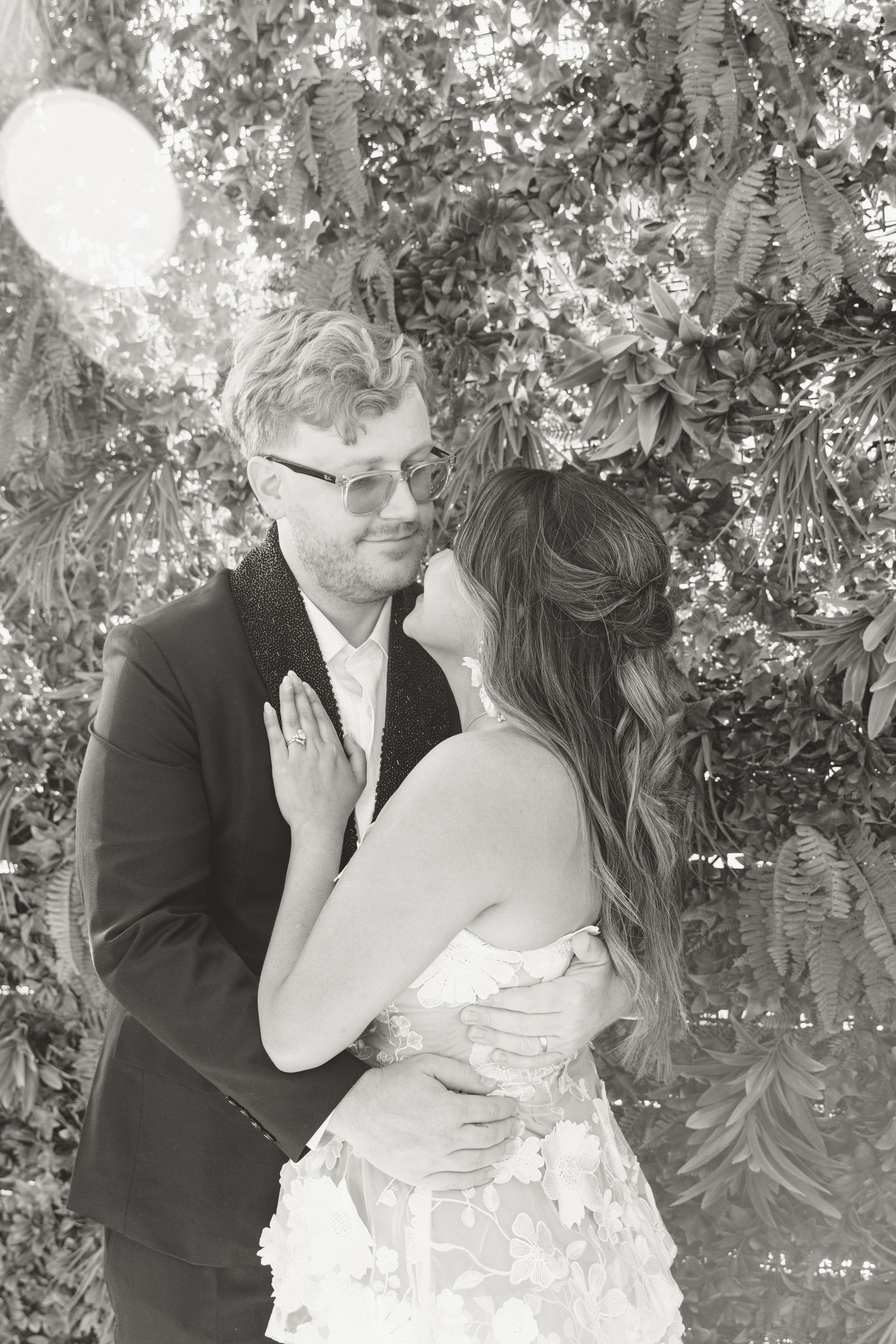 A black and white romantic portrait of newlyweds embracing among leafy foliage.