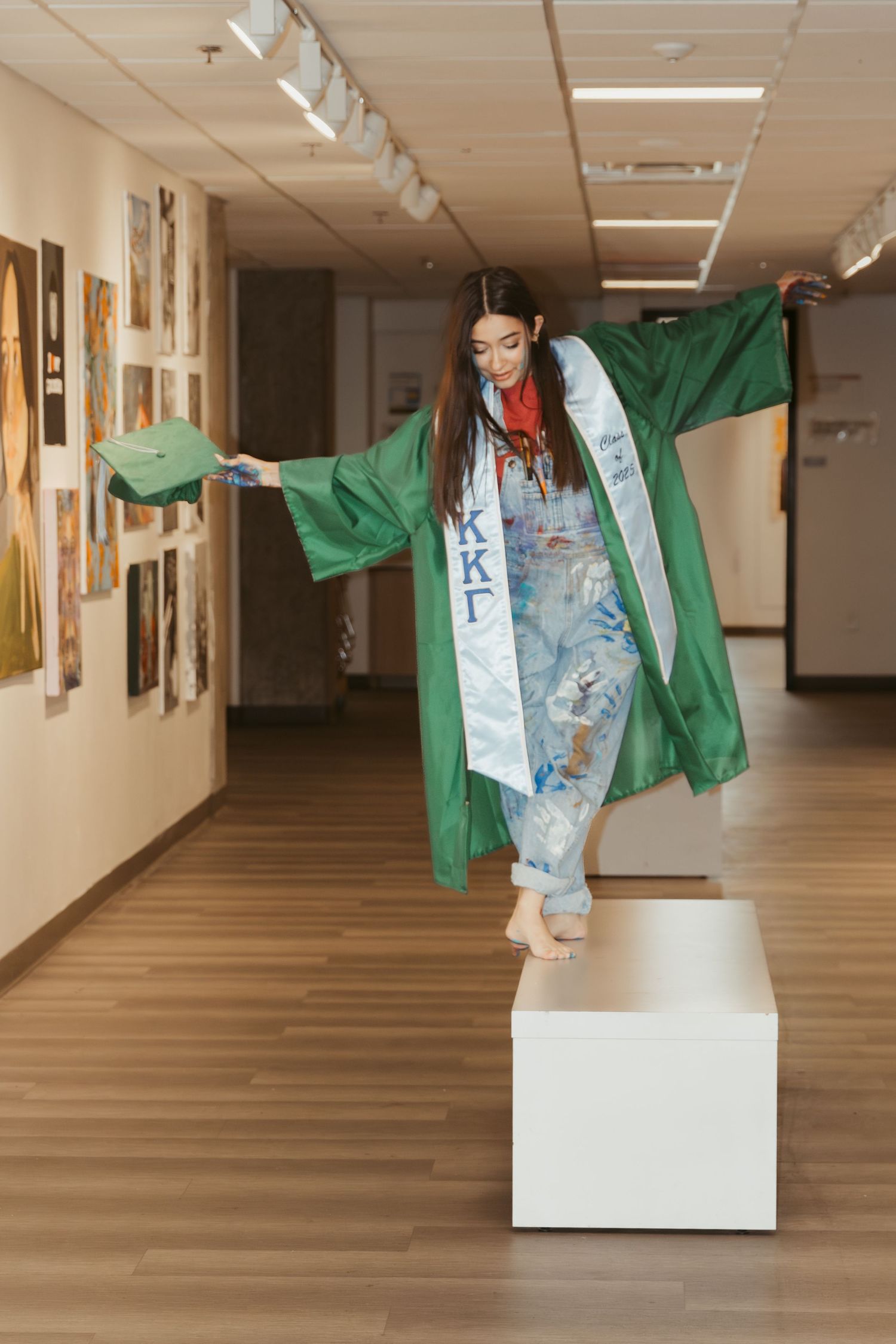 A graduate wearing a green gown and decorative overalls balances on a white pedestal in a hallway.