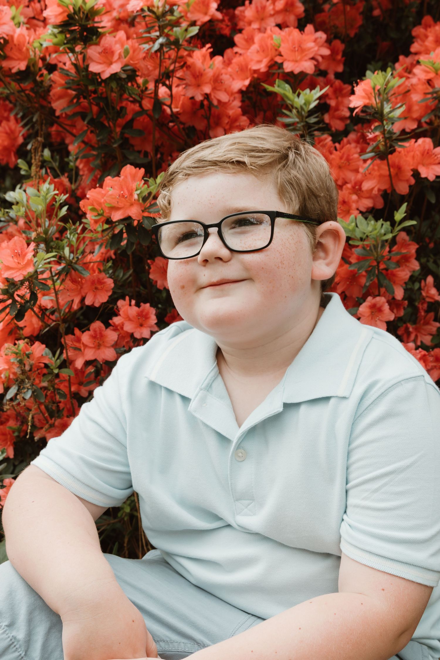 Child wearing glasses sits with bright coral azalea flowers blooming in the background.