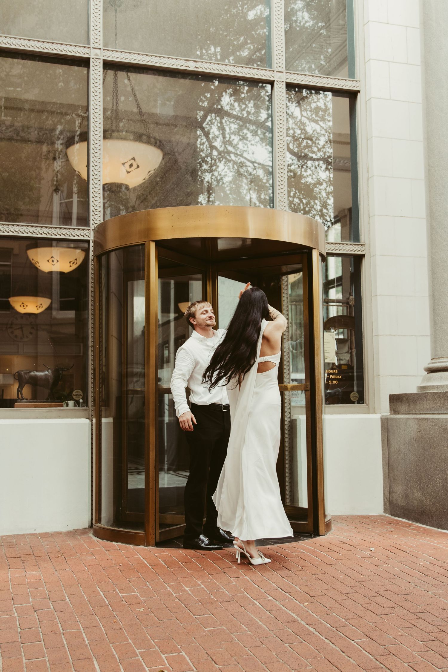 A couple in formal attire share a romantic moment in a revolving glass door entrance to an elegant building.