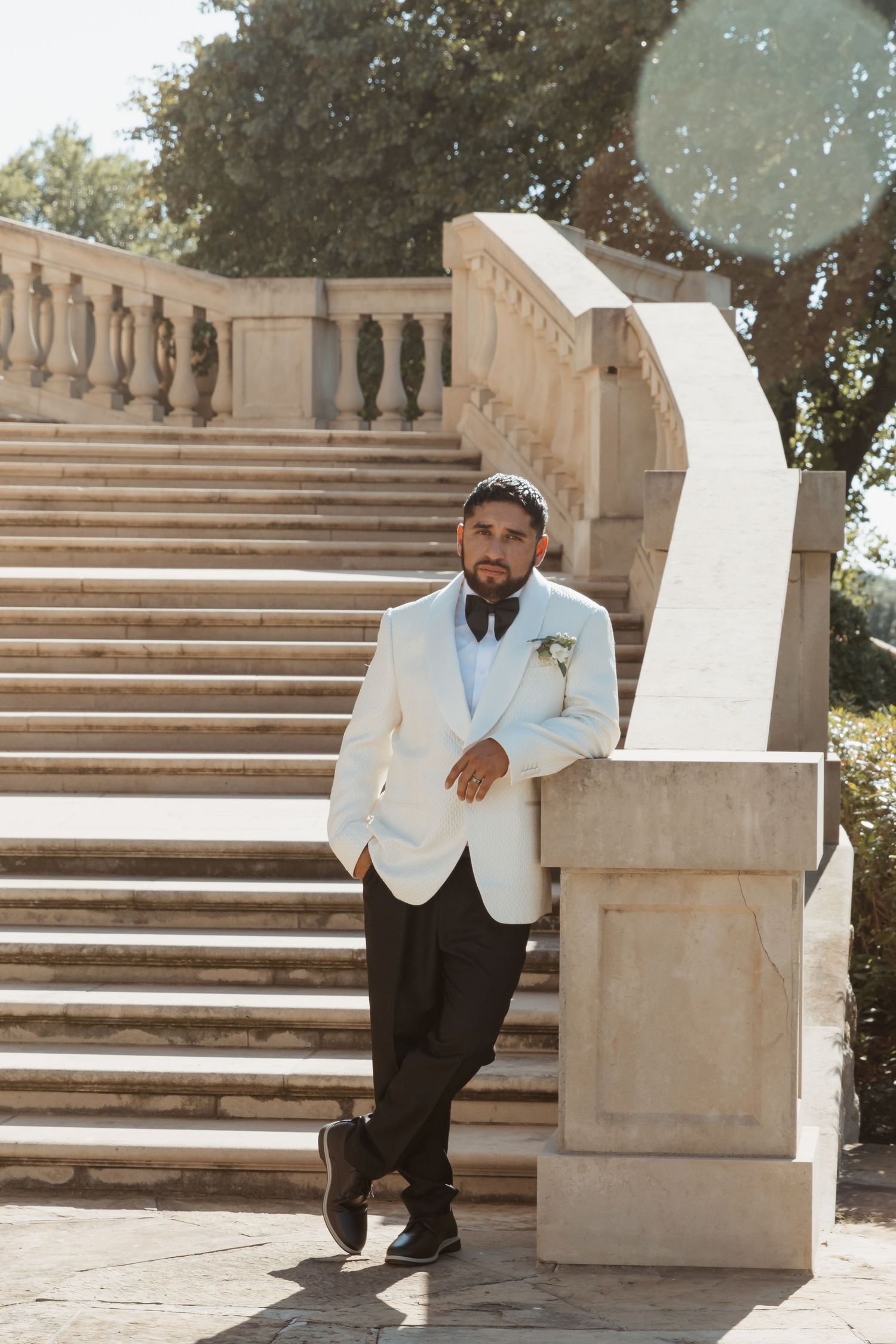 A person in a white tuxedo jacket and black pants sits elegantly on stone steps in an outdoor setting.