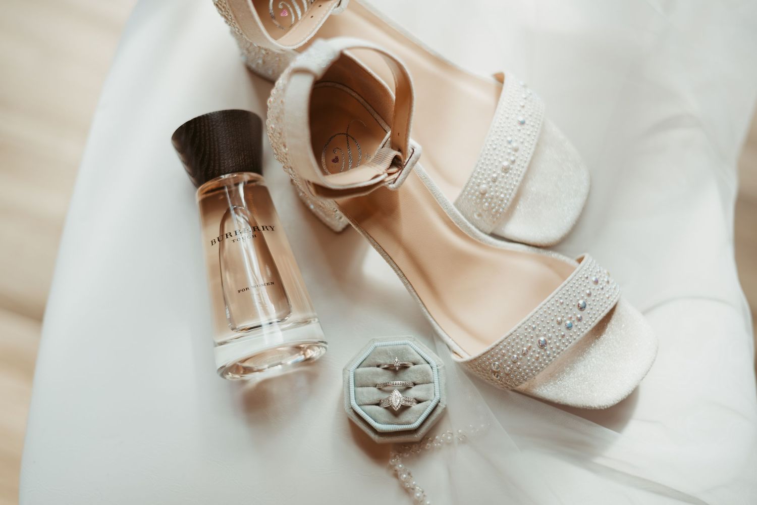 Elegant bridal accessories including white heels, perfume bottles, and delicate jewelry arranged on light fabric. Wedding photographer DFW
