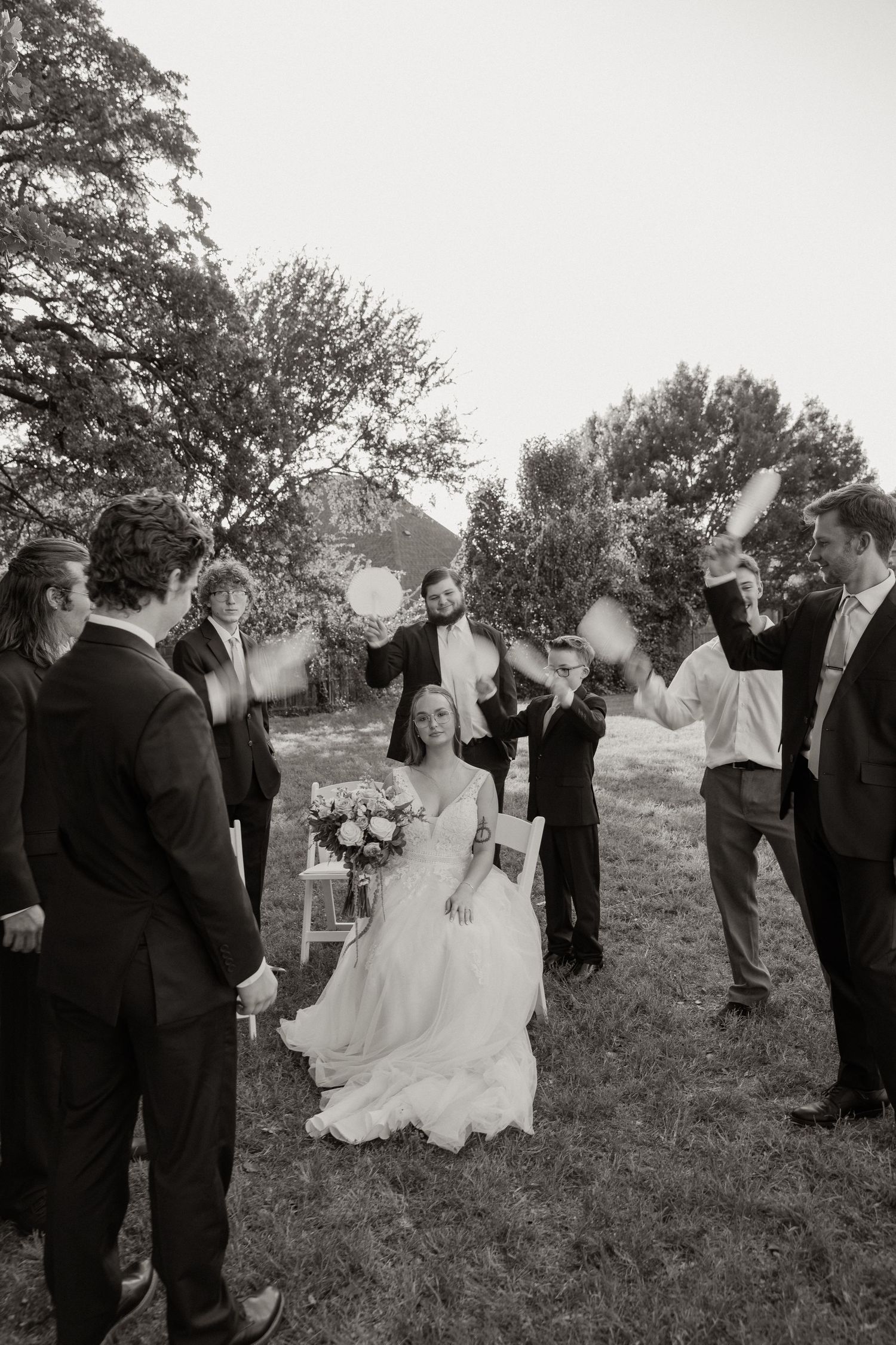 Bride seated in white gown as groomsmen celebrate around her during outdoor black and white wedding.