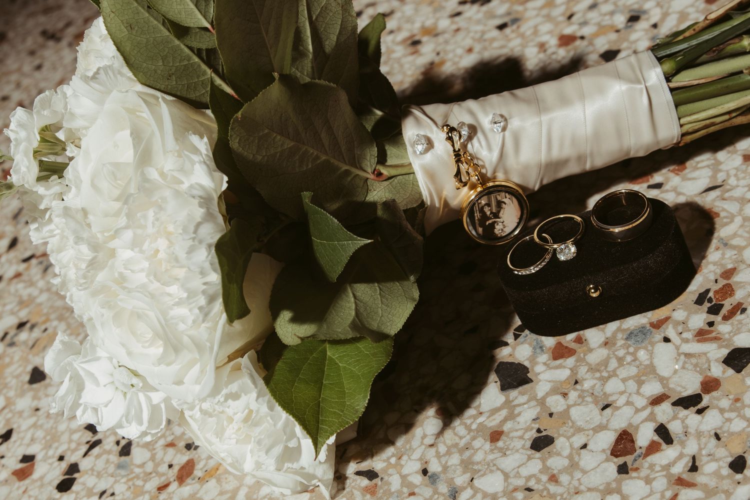 Black and white wedding rings nestled among white roses and greenery on a speckled surface. Wedding Photographer DFW