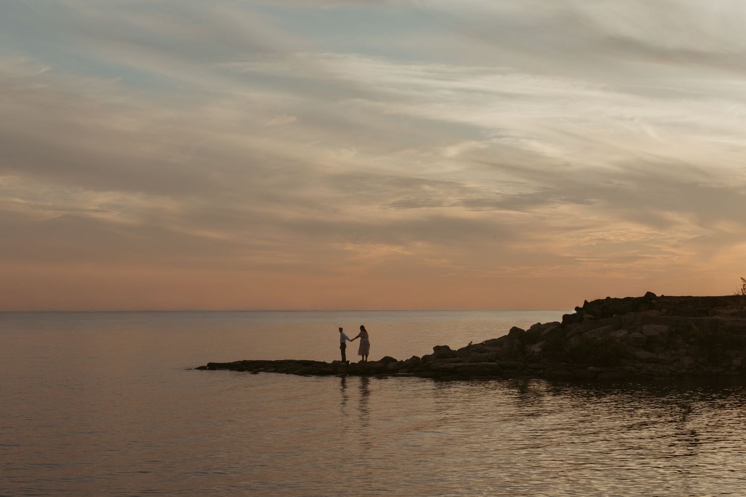Two silhouettes standing on rocky outcrop against sunset sky over tranquil water.
