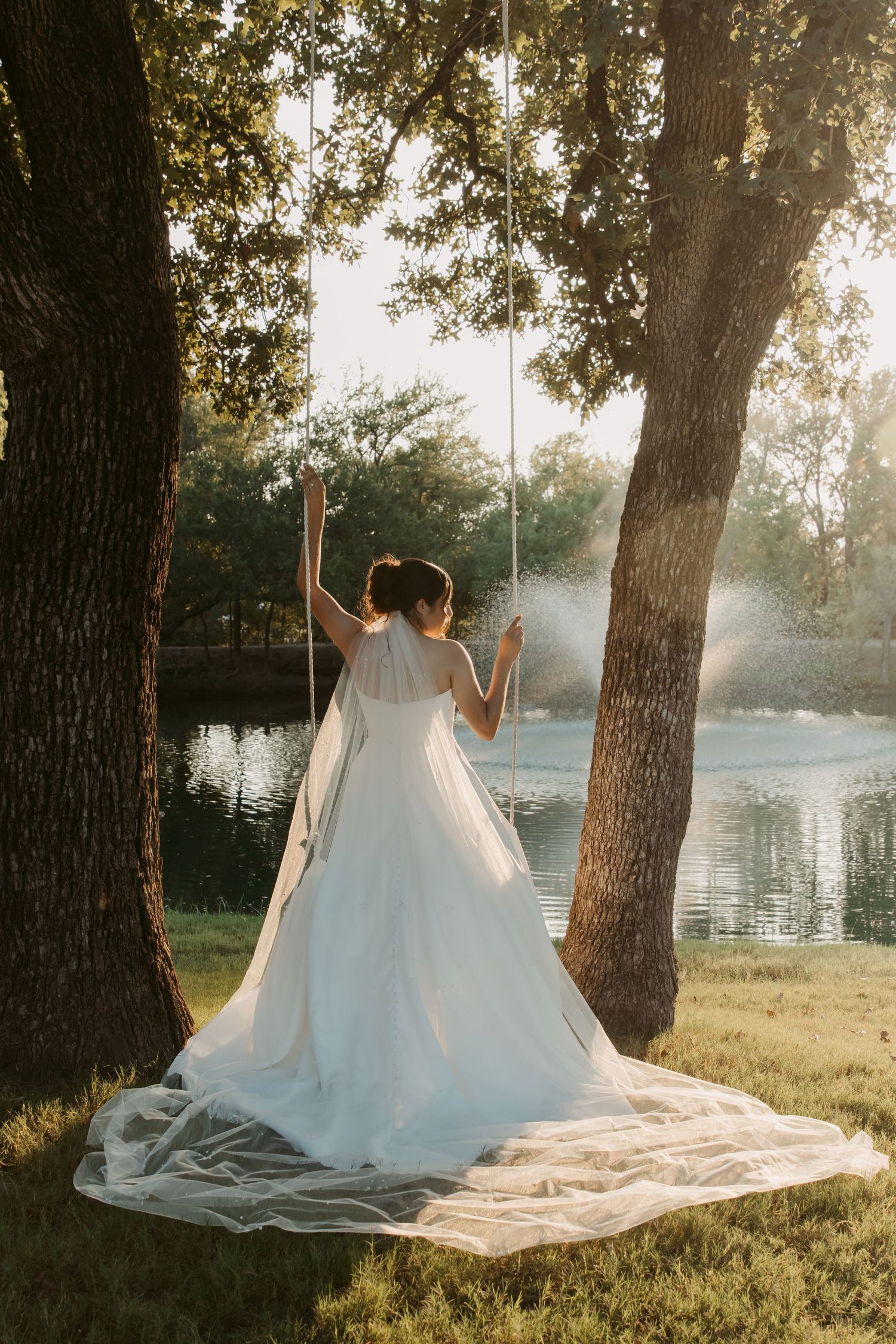 Flowing white wedding dress train spread across grass near a lake at sunset with trees in background.