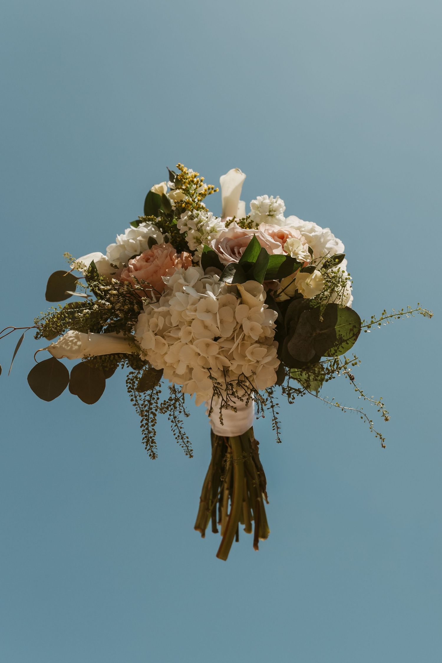 Elegant bridal bouquet with white hydrangeas, blush roses, and greenery held against a clear blue sky.