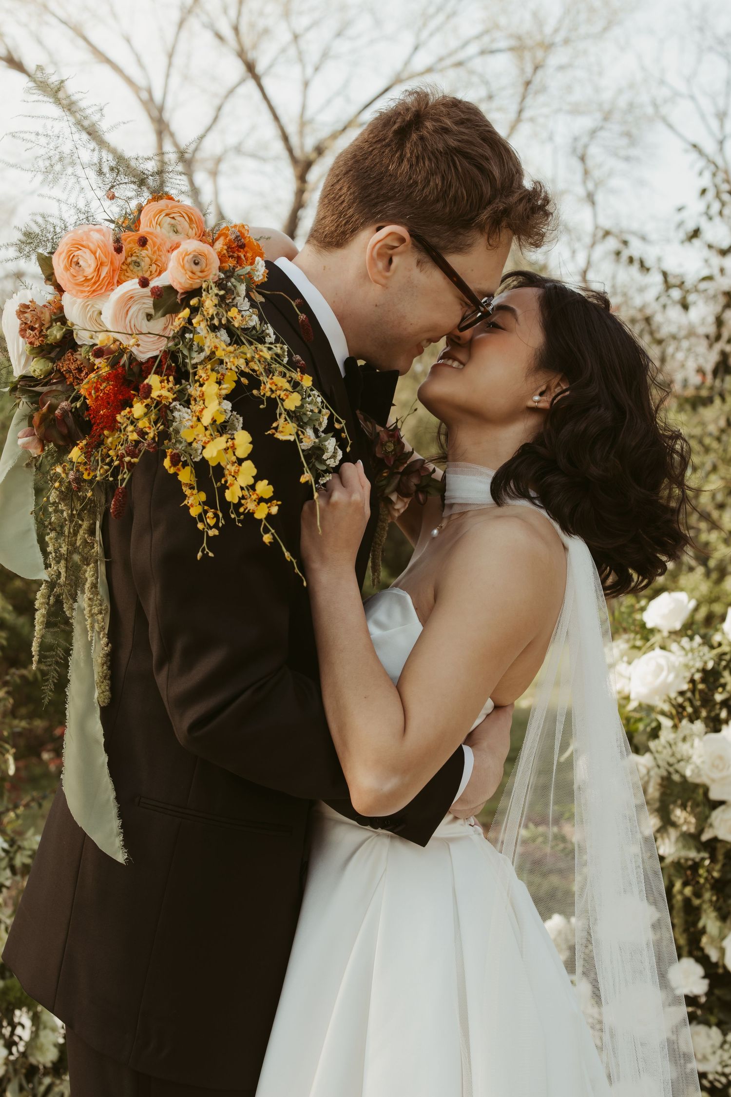 Bride and groom nearly kiss holding vibrant orange and yellow cascading bouquet at outdoor wedding ceremony.