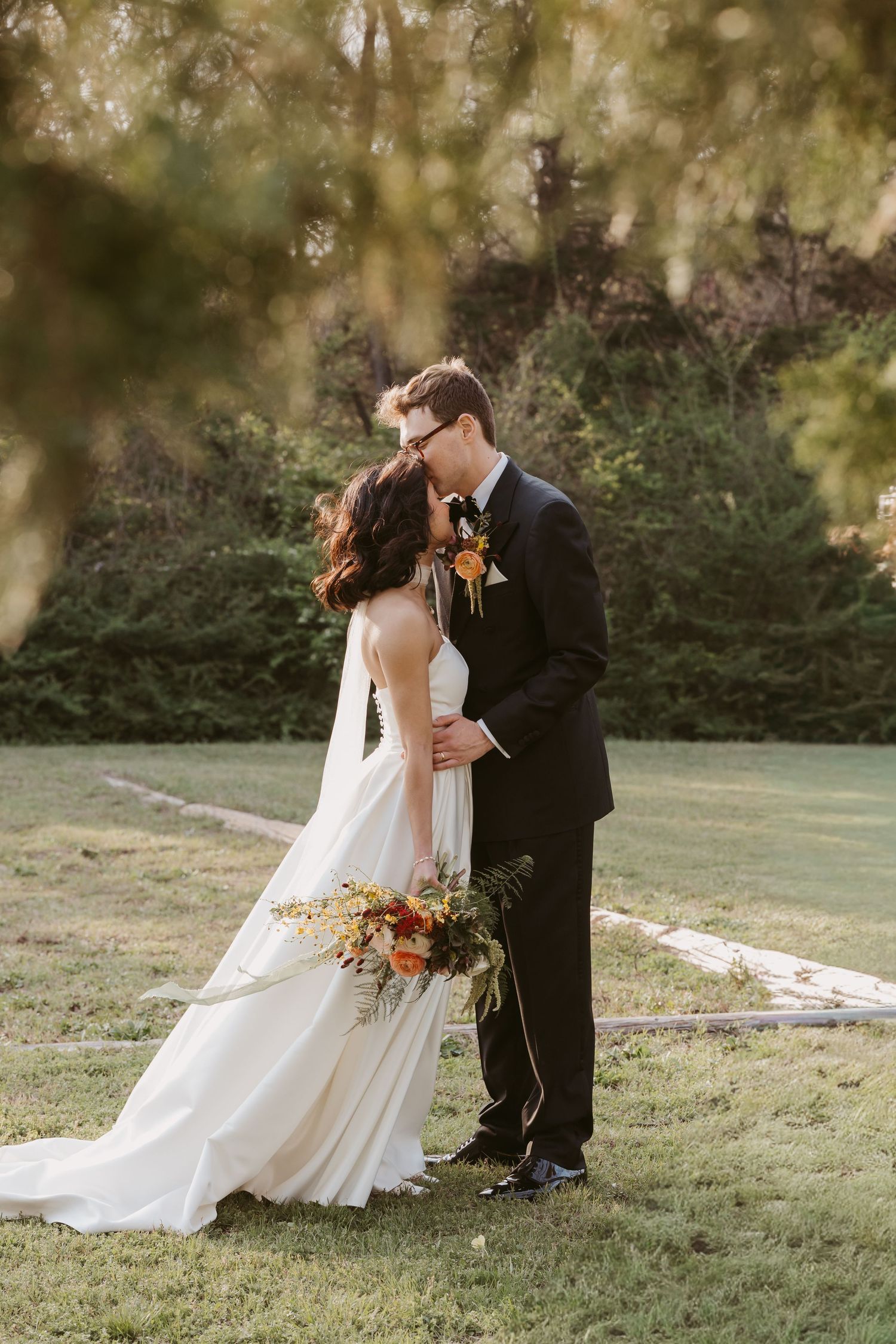 A bride and groom share a romantic kiss outdoors on their wedding day, surrounded by lush green trees and golden sunlight.