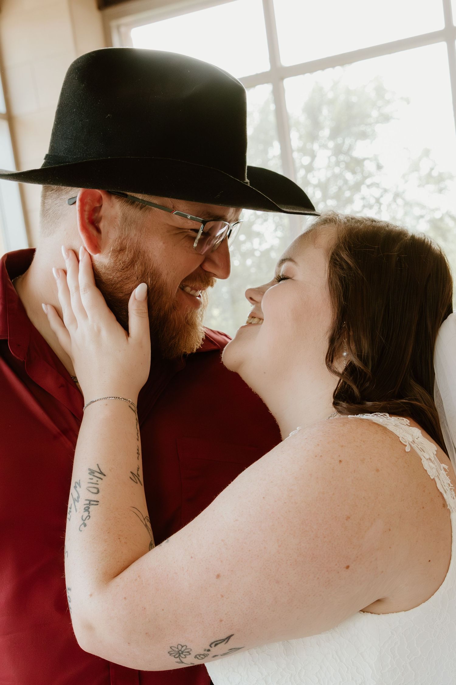 Intimate wedding portrait with burgundy shirt and black hat against window light.