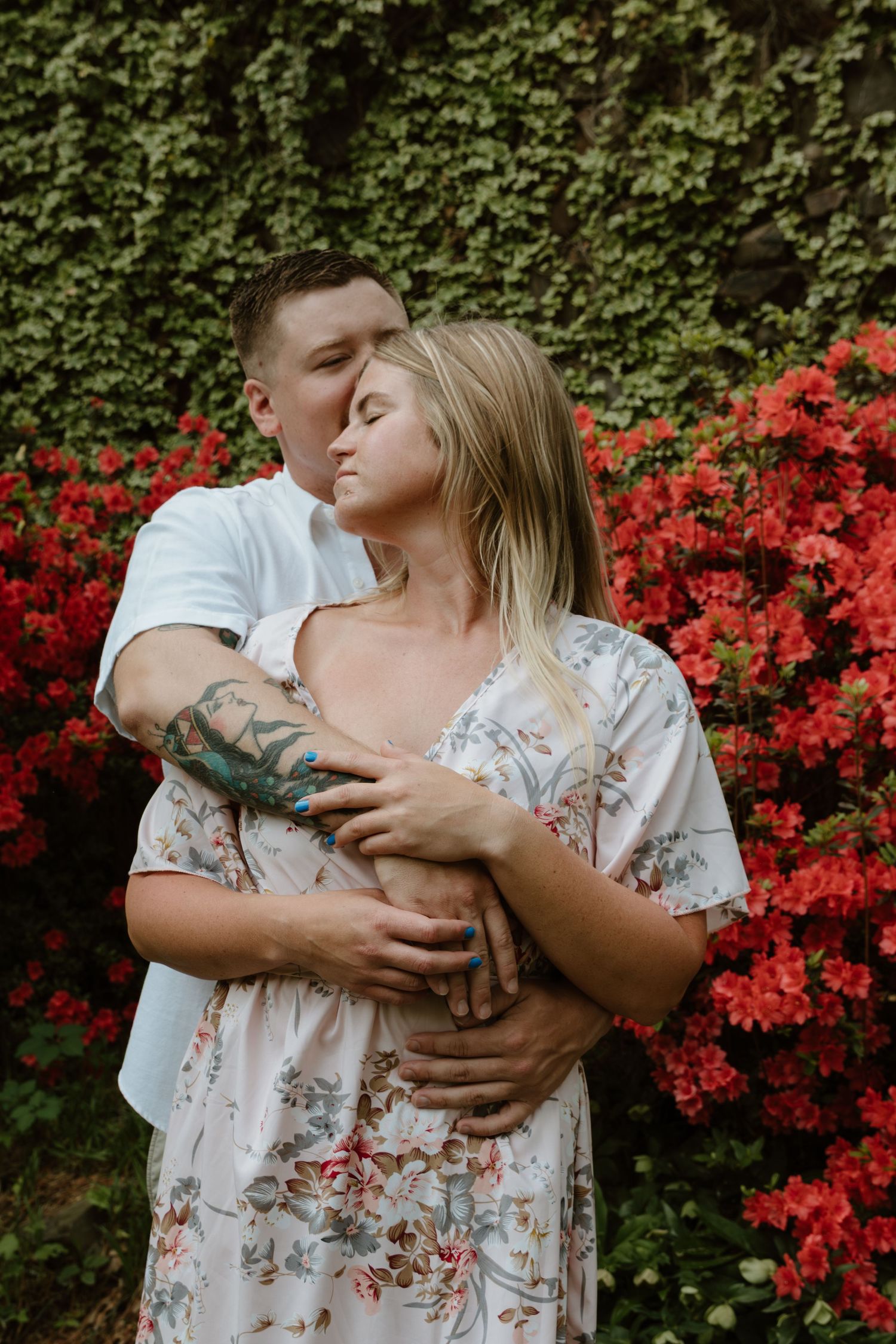 A romantic couple embracing against a backdrop of vibrant red azalea flowers and ivy-covered walls.