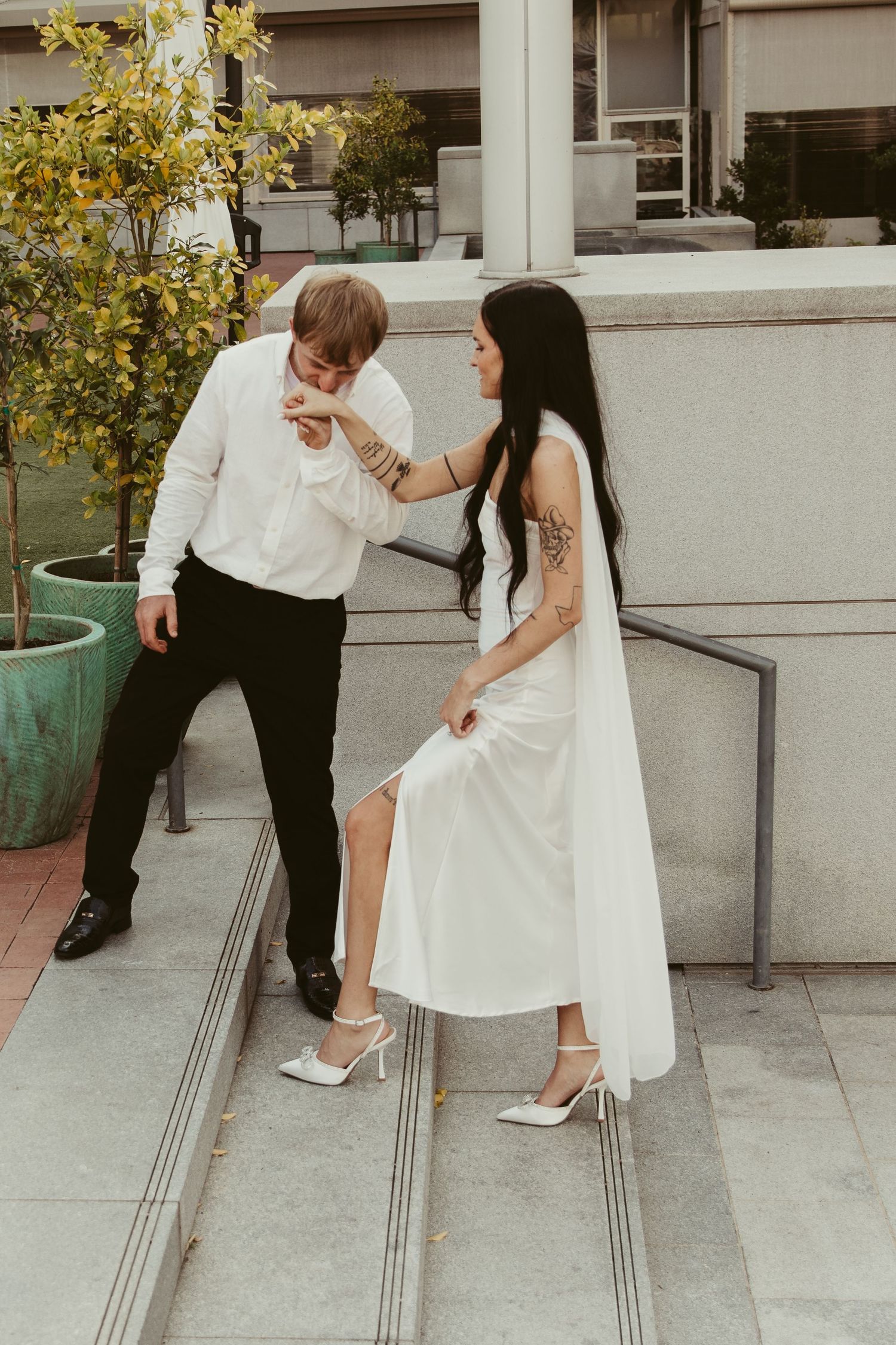 A couple in wedding attire shares a playful moment on outdoor steps beside a turquoise planter with yellow flowers.