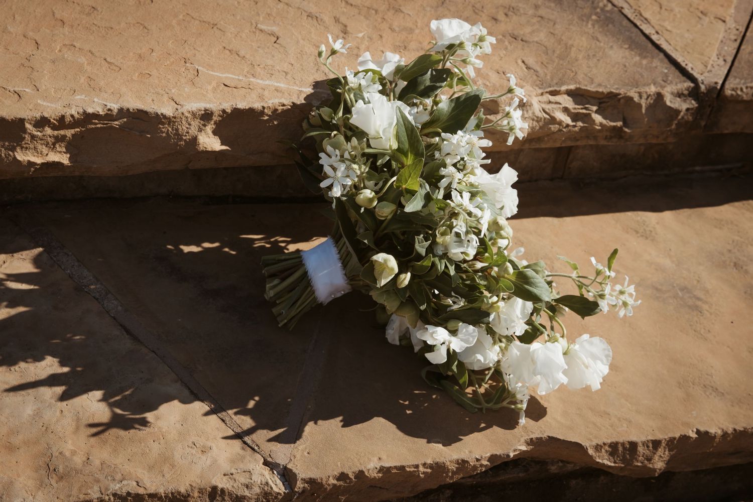 A white wedding bouquet with mixed flowers and greenery lies on a rustic wooden surface casting soft shadows.