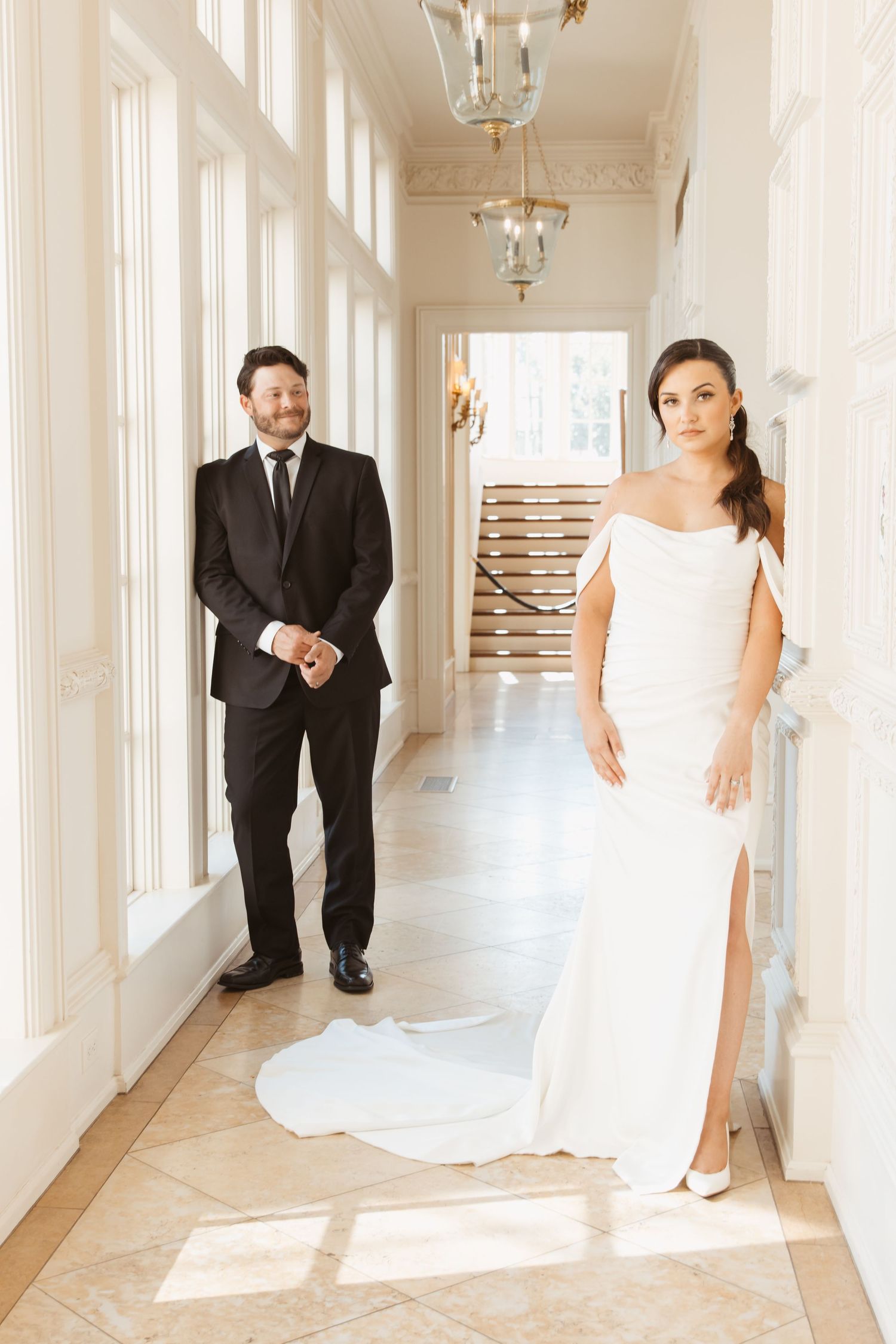 A wedding couple poses in a bright white hallway with elegant pendant lighting and tall windows.
