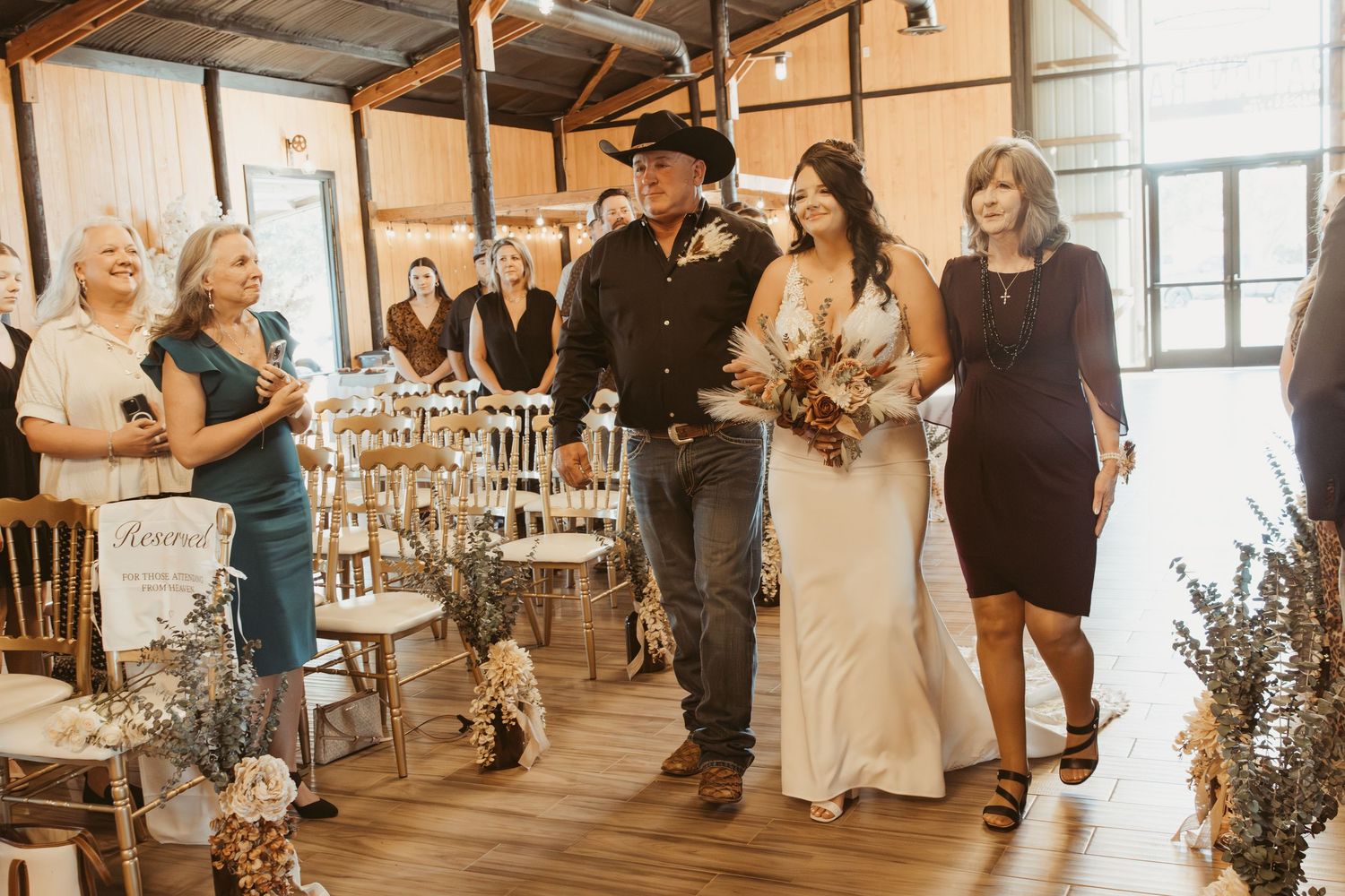 A western-themed wedding ceremony processional in a rustic indoor venue with wooden floors and natural light. Wedding photographer DFW