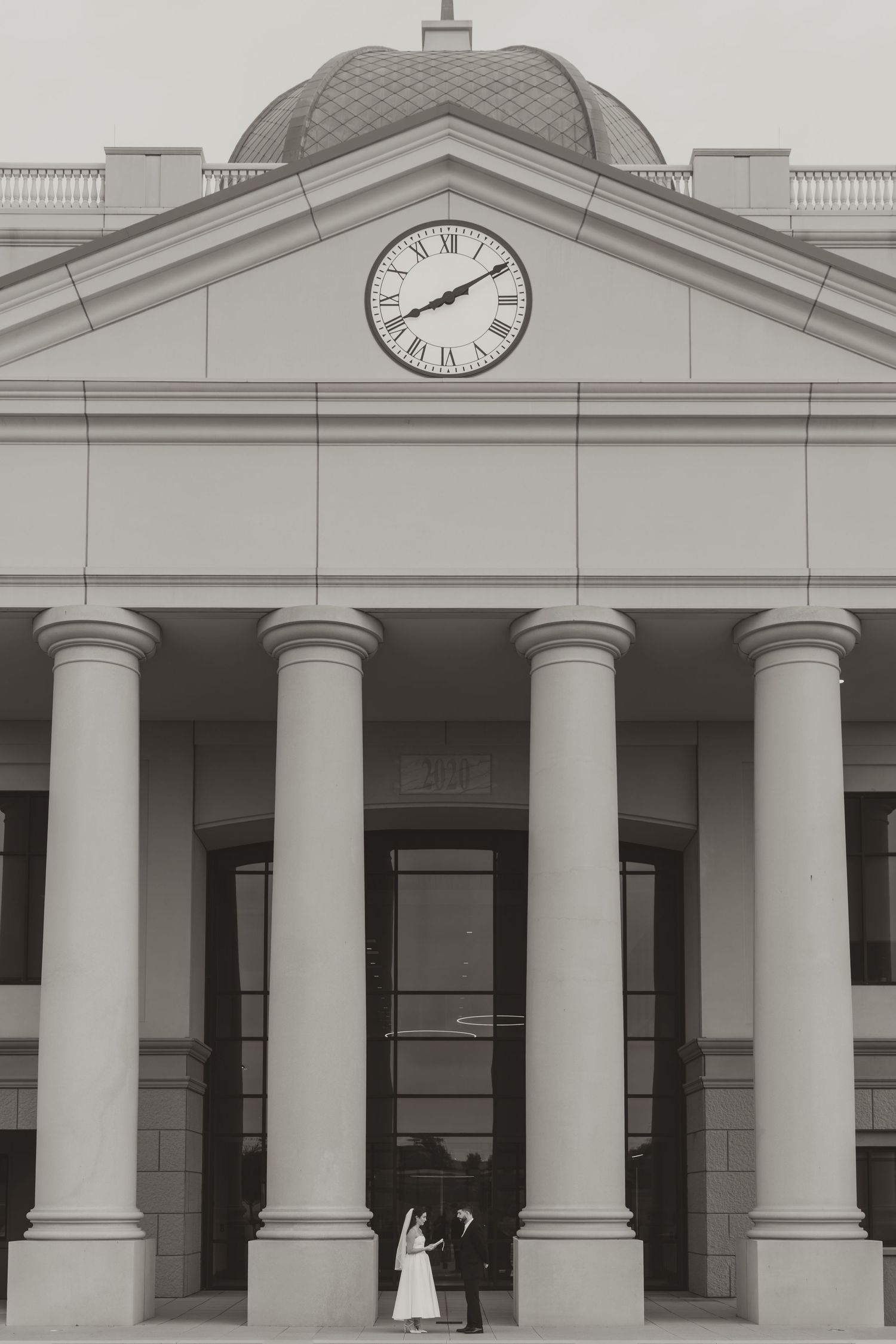 A black and white architectural photo of a courthouse with columns and a clock, with a couple standing below. Wedding Photographer Fort Wort