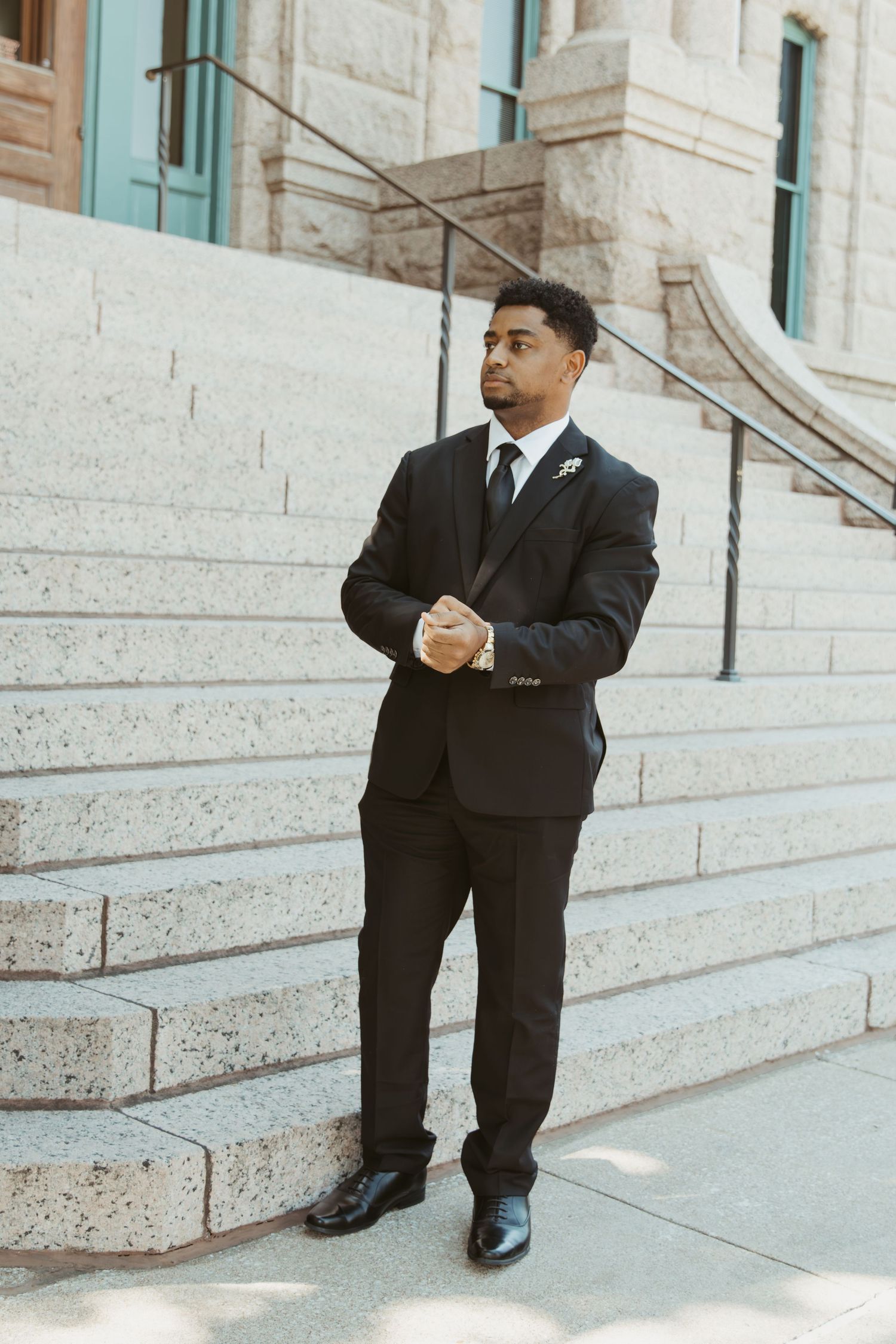 A person in a dark suit sits thoughtfully on stone steps outside a building with turquoise accents. Wedding Photographer DFW