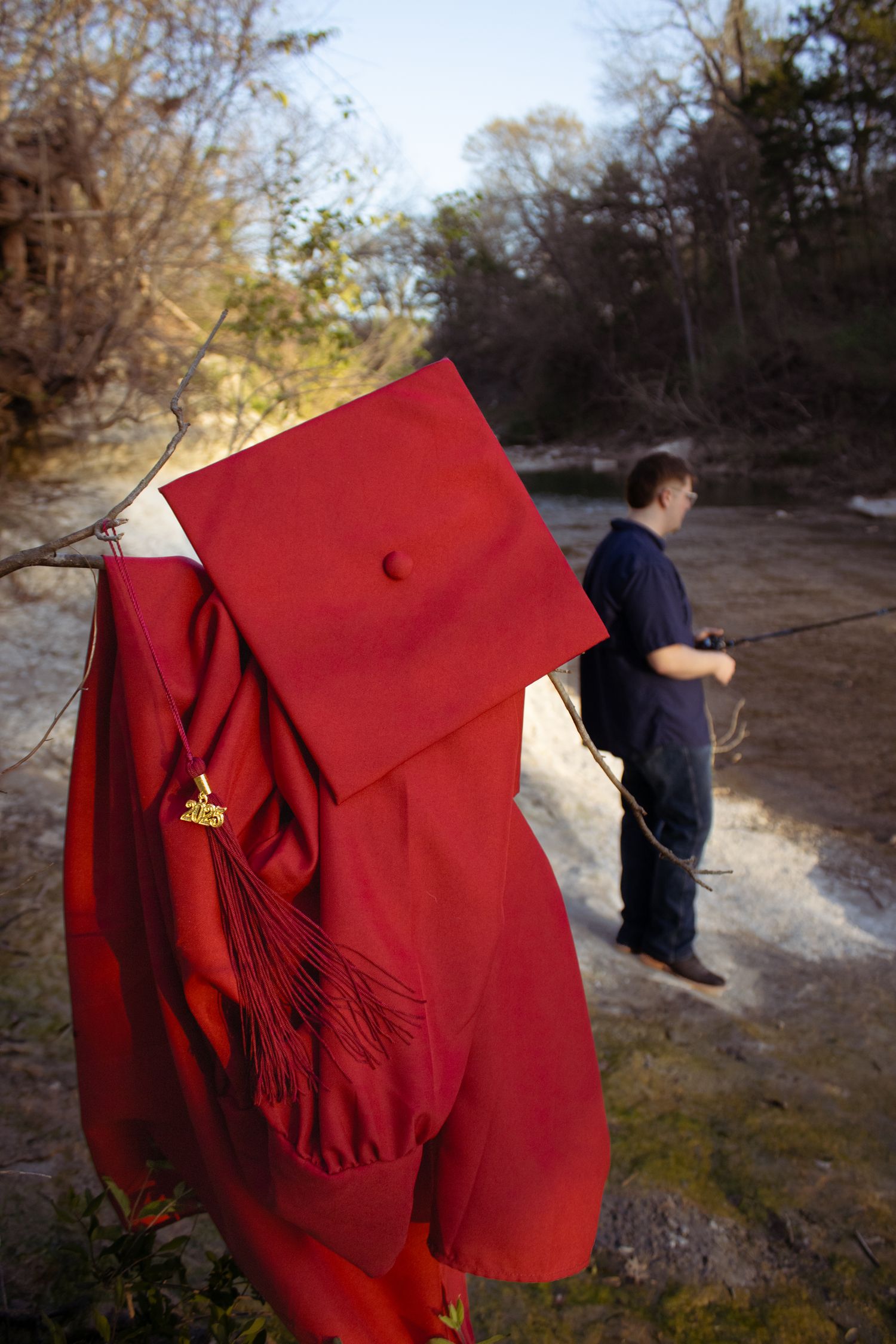 A red graduation cap and gown hanging outdoors with someone fishing by a river in the background.