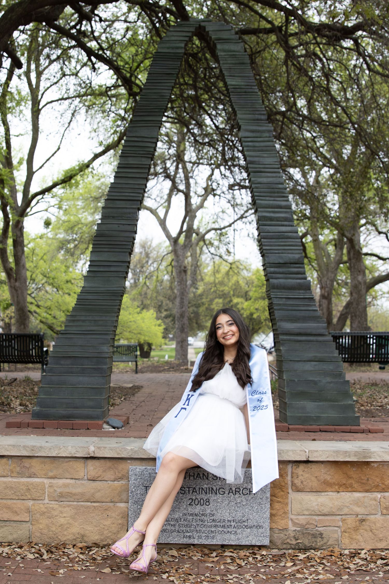 Graduate in white dress and blue sash sitting beneath a large metal arch structure outdoors.