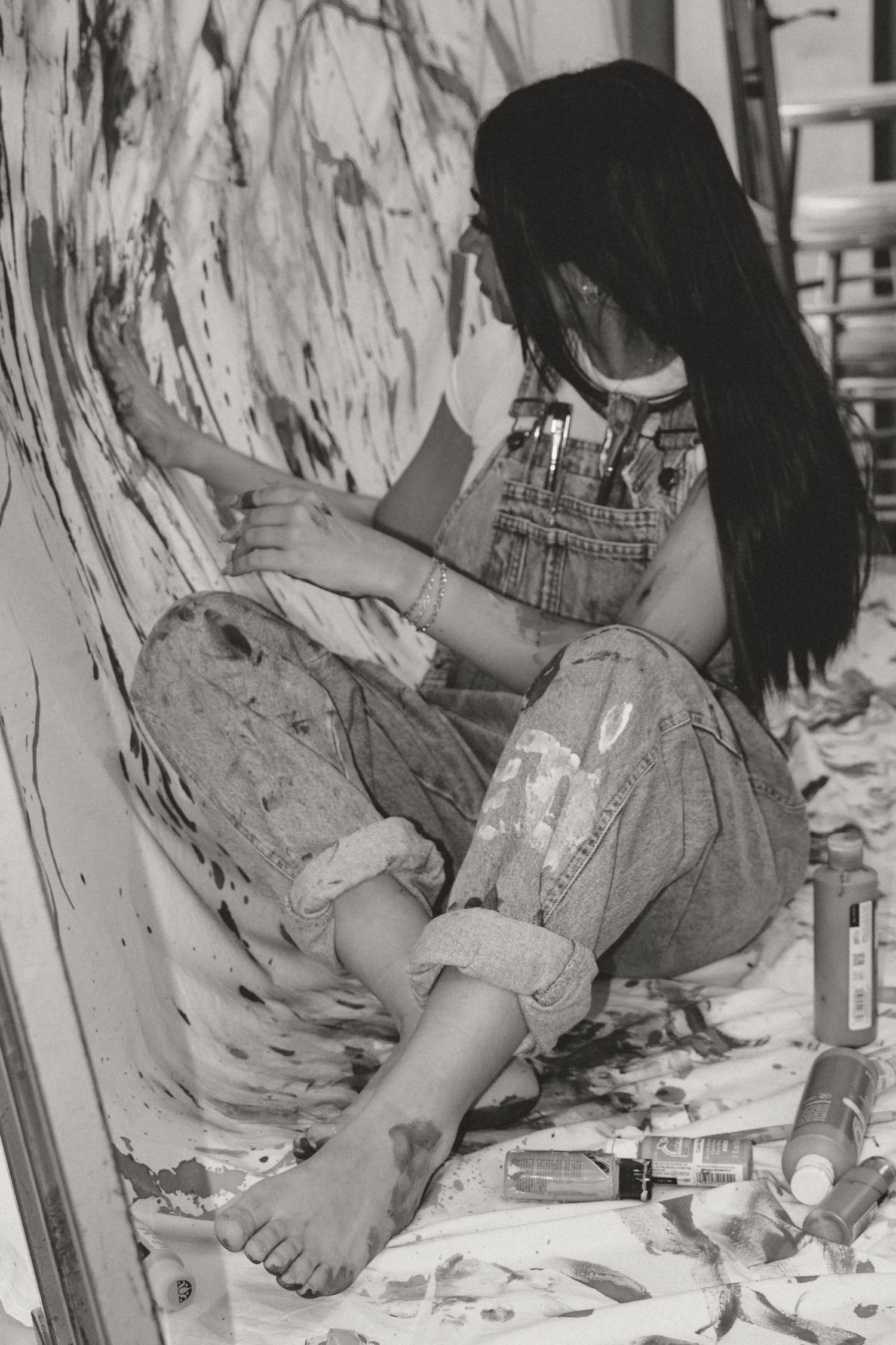 Black and white photo of an artist in overalls sitting on the floor painting a large mural with paint supplies scattered around.