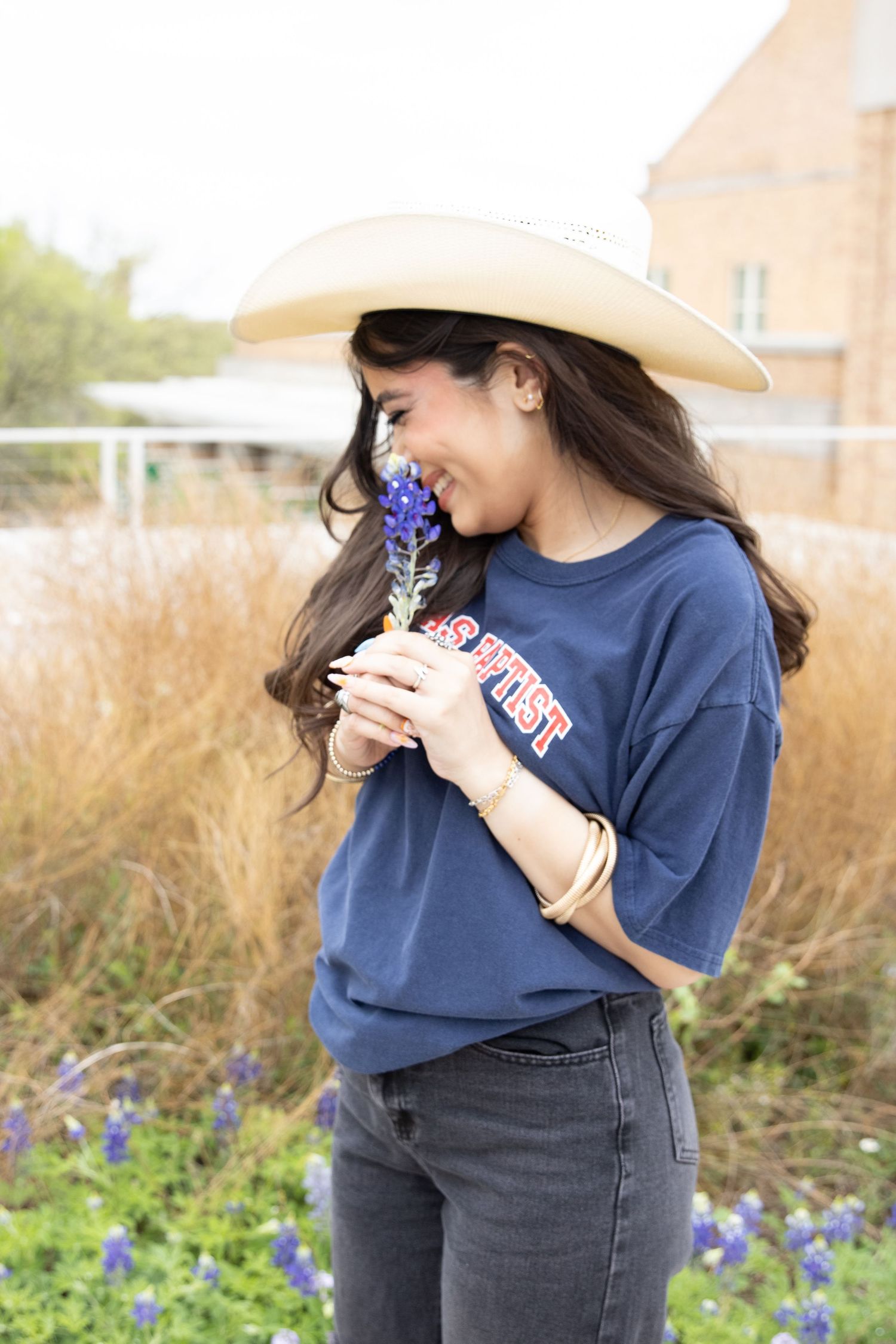 Someone wearing a white cowboy hat and navy shirt smells bluebonnets while smiling in a wildflower field.