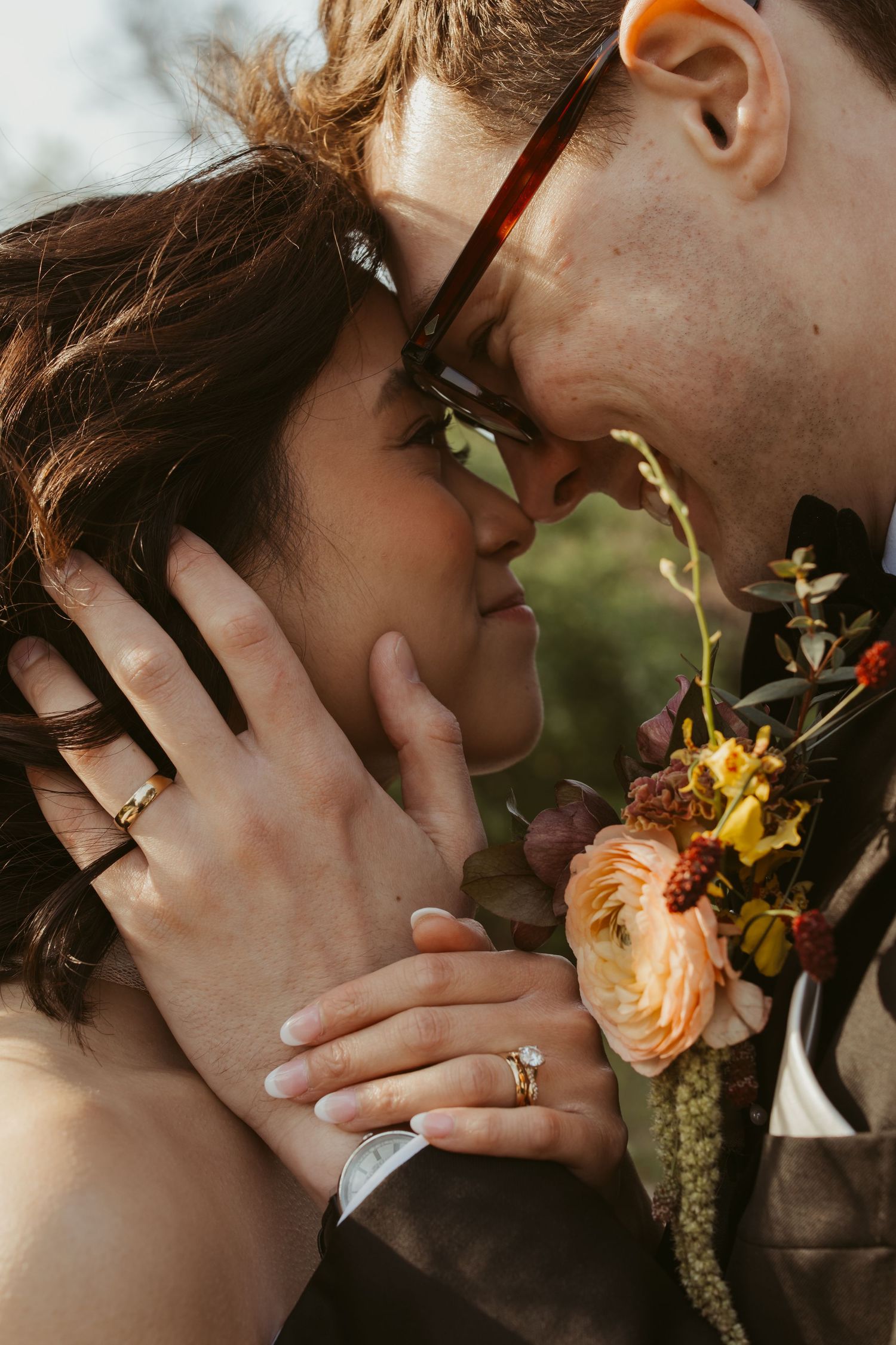 Close-up of bride and groom touching foreheads, showing wedding rings and floral boutonniere.