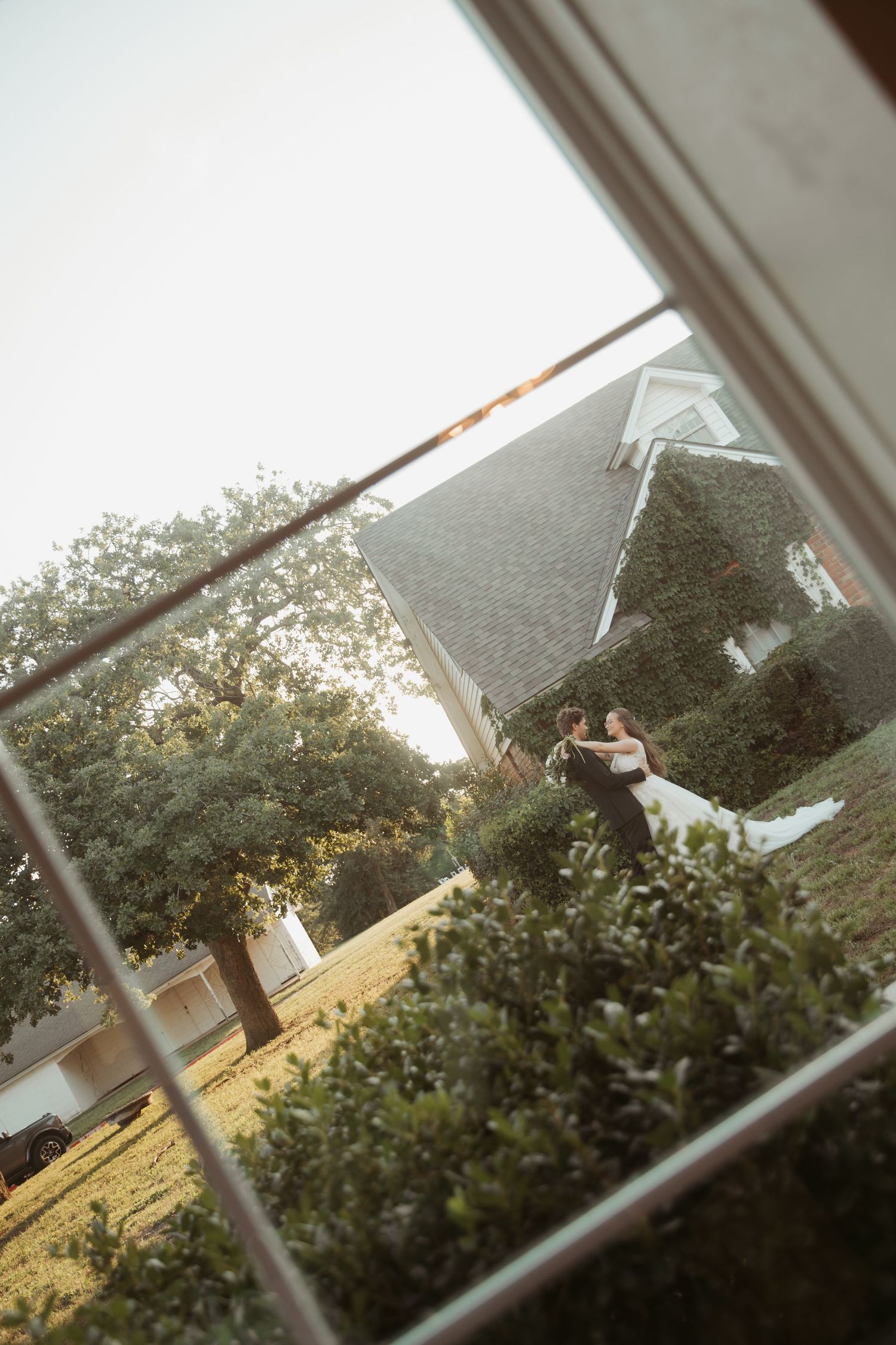 Bride and groom embracing outdoors near an ivy-covered church, photographed through a white window frame.