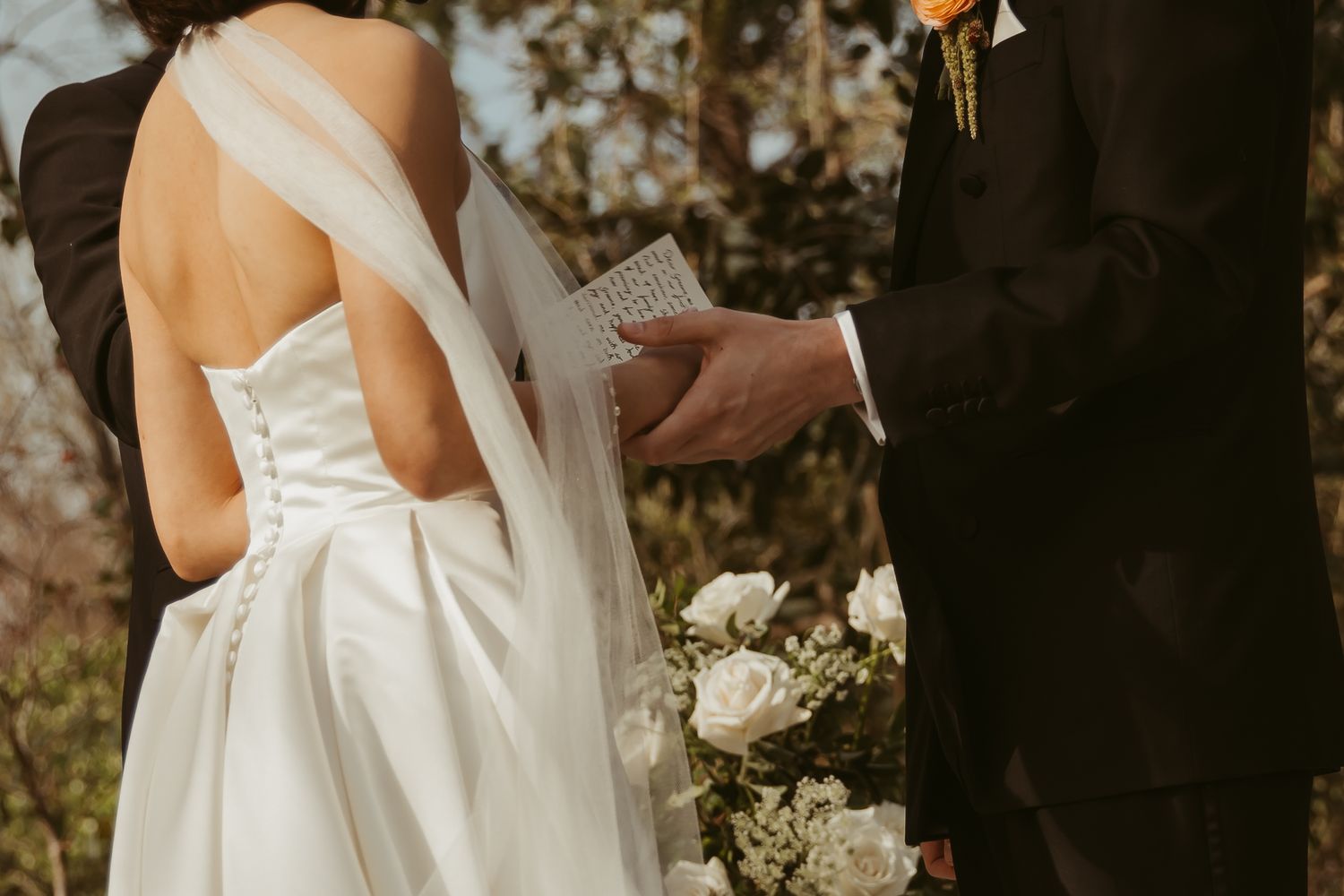 Bride in elegant backless white gown with veil stands beside groom in dark suit during outdoor wedding ceremony.