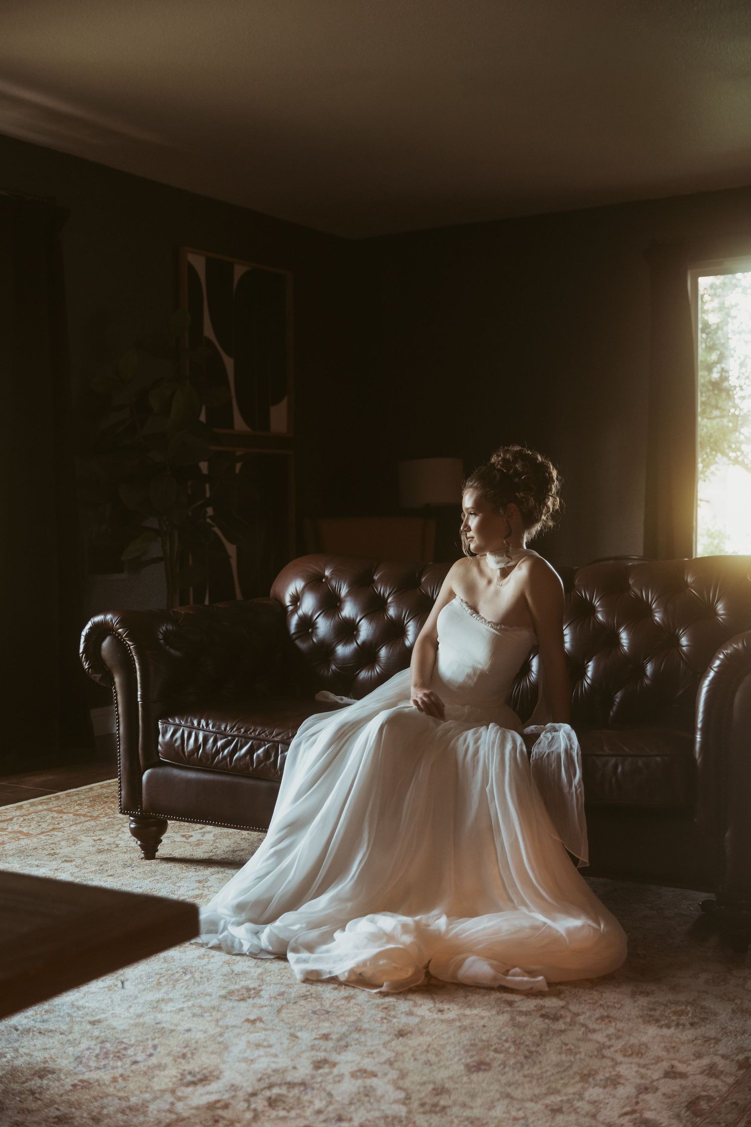 Bride in white gown sitting on leather Chesterfield sofa in dimly lit room with warm window light.