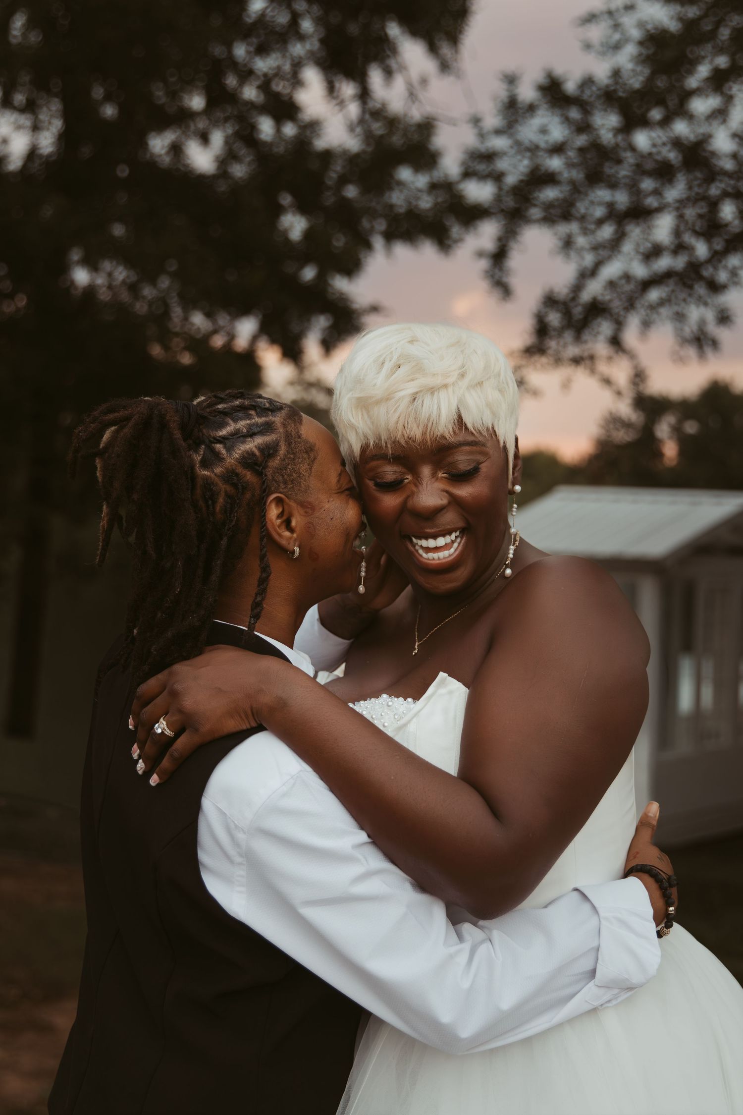 Two people share an intimate embrace outdoors during sunset wearing white clothing.