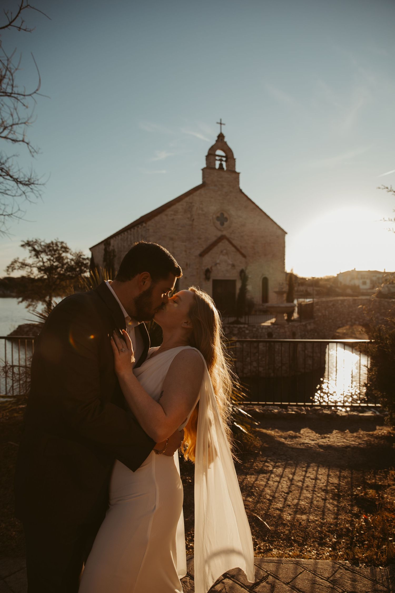 Newlyweds share a romantic kiss at sunset in front of a rustic stone church with mountains in the background.