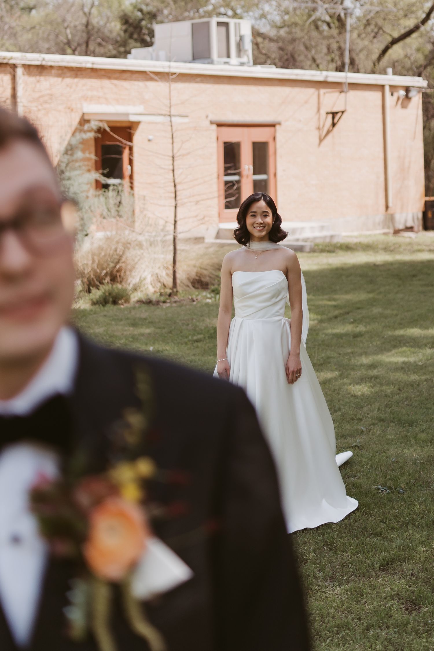 Bride in strapless white gown smiling at groom in black tuxedo during outdoor wedding at brick venue.