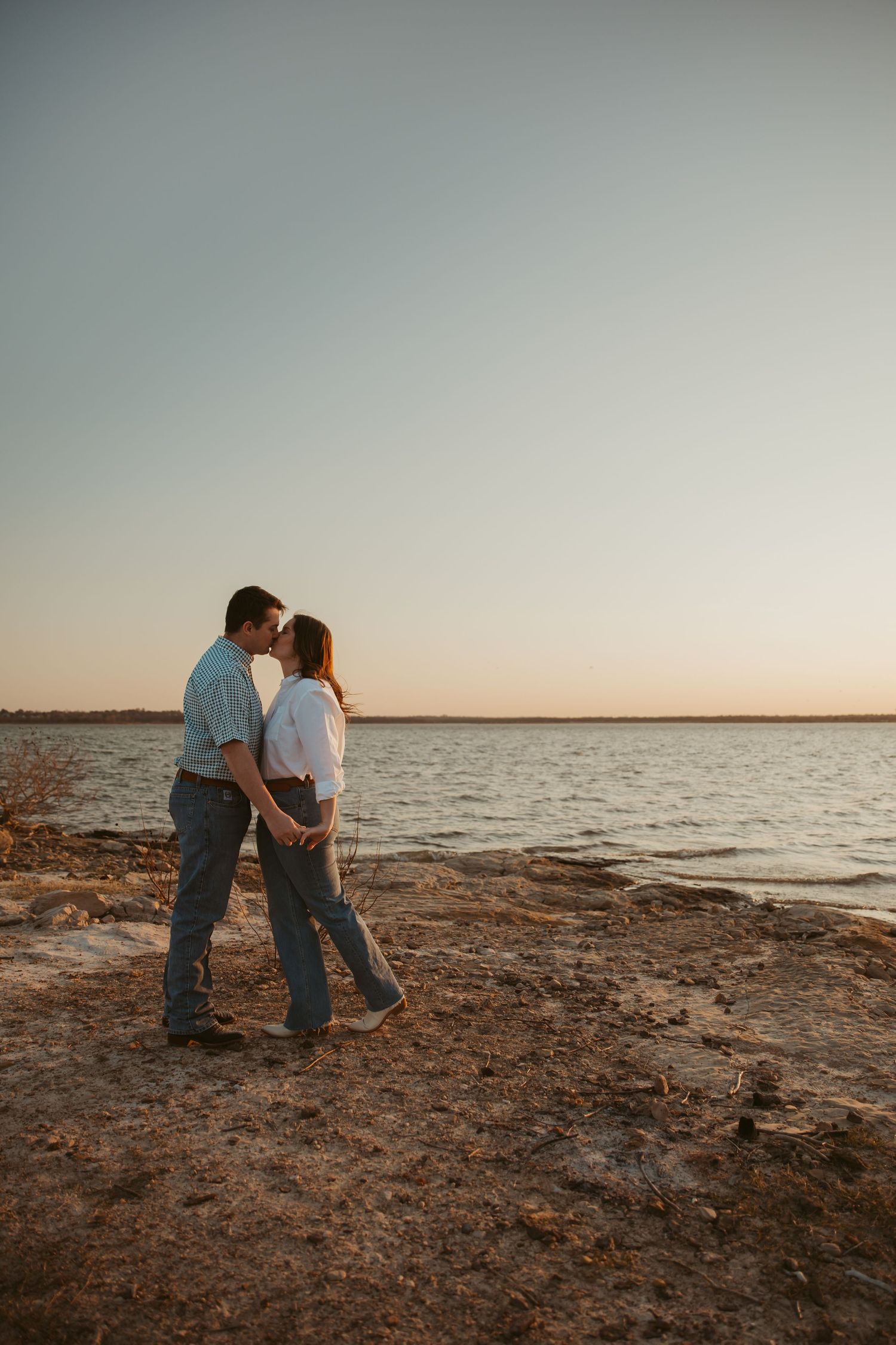 Couple sharing a romantic kiss at sunset by a serene lakeside, dressed casually in jeans and light tops.