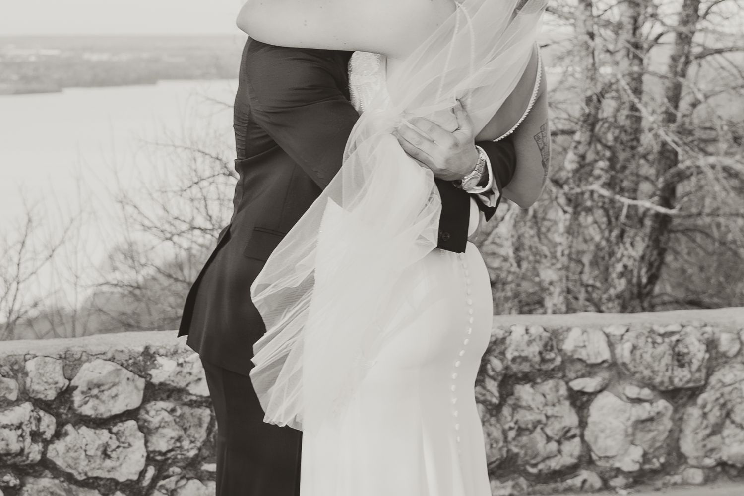 Black and white wedding photograph showing a close embrace between a couple against a stone wall overlooking a scenic view.